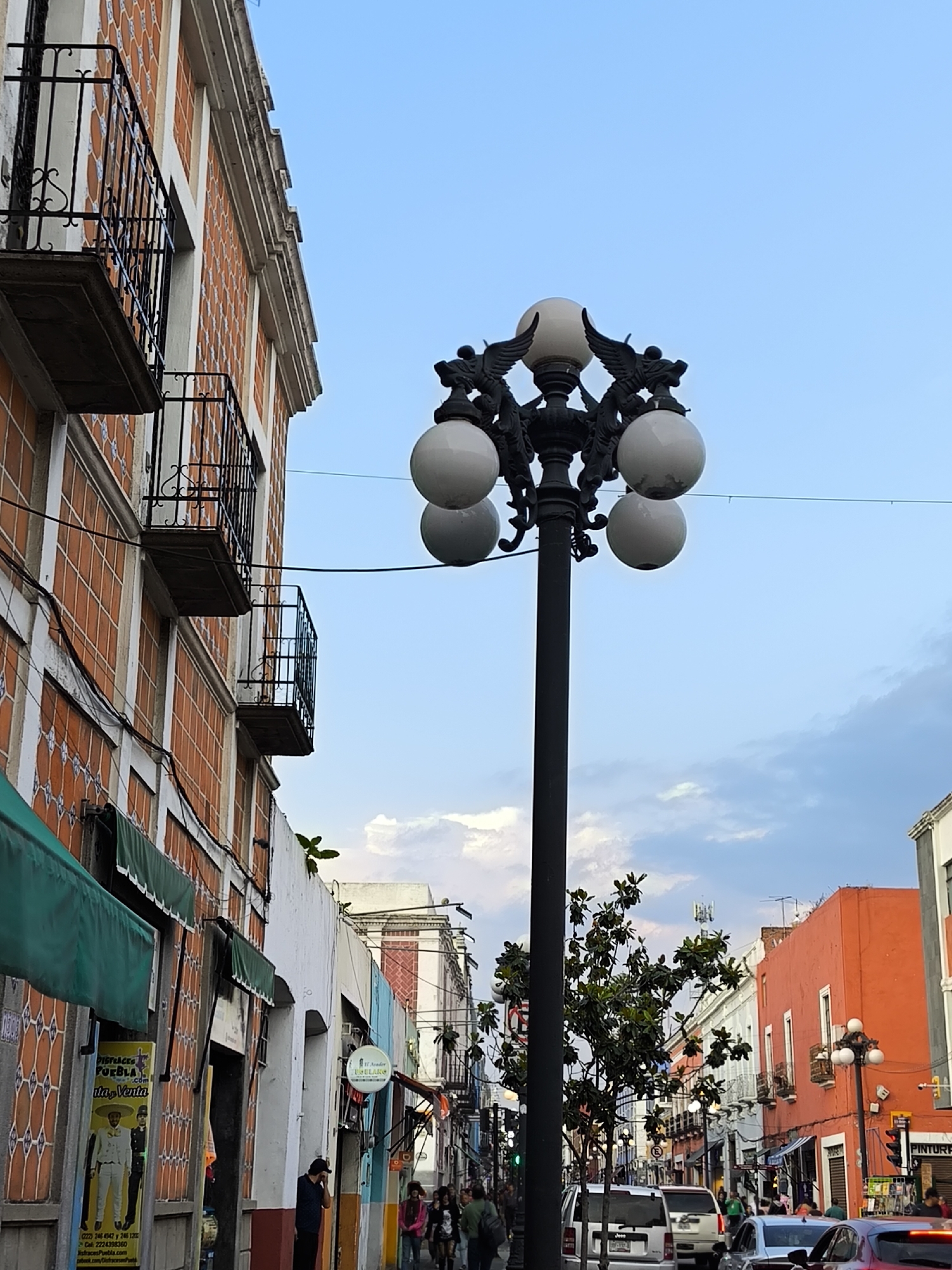 The photograph depicts a street scene in the city of Puebla, Mexico, characterized by historical architecture and street activity. In the foreground is a wrought-iron street lamp, adorned with spherical white globes and decorative winged figures. The lamp post rises against a backdrop of a partly cloudy sky that transitions from a pale blue to a slightly darker hue.
To the left, the facade of a multi-story building is visible, featuring orange tiled walls and ornate iron balconies. The lower levels of the building appear to have shops with green awnings. A poster for "Disfraces Puebla" can be seen, showing a man dressed in traditional Mexican attire, advertising costumes for sale.
Moving down the street, a mix of pedestrians and vehicles can be observed, indicating a moderately busy urban environment. There's a variety of architectural styles, including buildings painted in bright colors like orange, interspersed with more traditional white facades. Other street lamps line the street, and there is a traffic light visible in the distance.