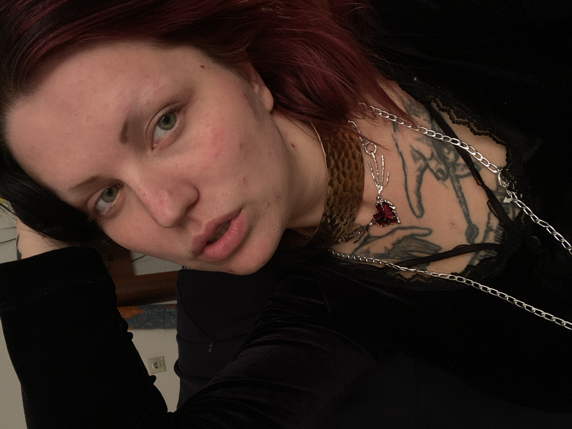 A close-up, angled shot depicts a fair-skinned individual with light green eyes and reddish-brown hair. Their hair is predominantly black on one side and reddish-brown on the other. The person is wearing black clothing with lace trim at the neckline. A prominent tattoo is visible on the upper chest and neck. Two necklaces adorn the individual: one is a wide, textured gold collar, and the other is a long silver chain necklace with a pendant of a hand holding a red gem. The background includes a glimpse of a wooden headboard and a light-colored wall with an electrical outlet. No evidence indicates the location is in Kunje, Montenegro.