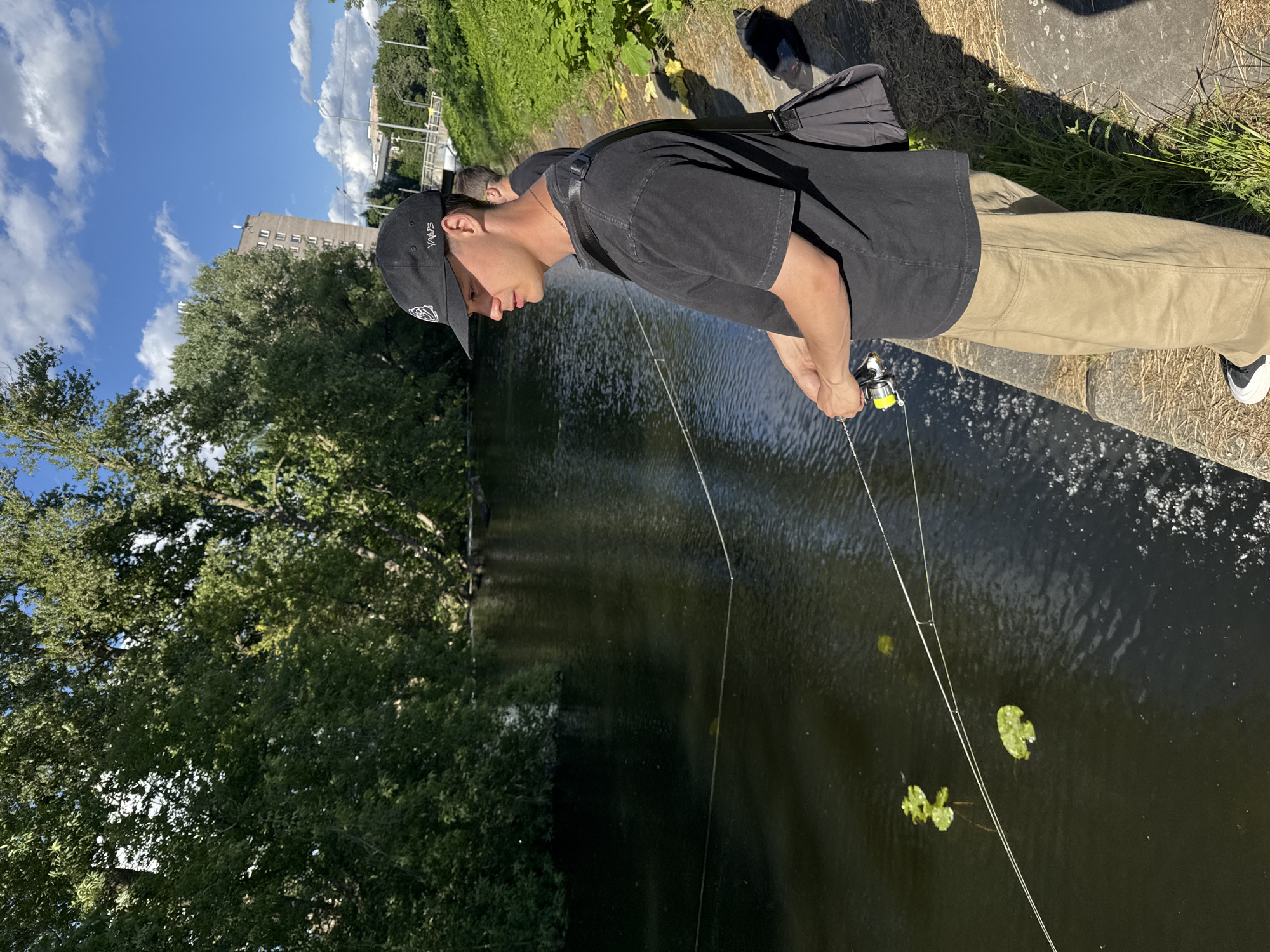 In Saint Petersburg, Russia, a young Caucasian male wearing a black baseball cap, a black t-shirt, and khaki pants stands on the paved bank of a dark, slow-moving river or canal. He is holding a fishing rod with his right hand, and his gaze is directed towards the water, suggesting he is actively fishing. A black shoulder bag is slung across his chest. In the background, lush green trees line the opposite bank, and a multi-story building is visible further in the distance. The sky is blue with scattered white clouds. Another person, partially visible, is standing behind the primary subject. The river has a few scattered lily pads on its surface.