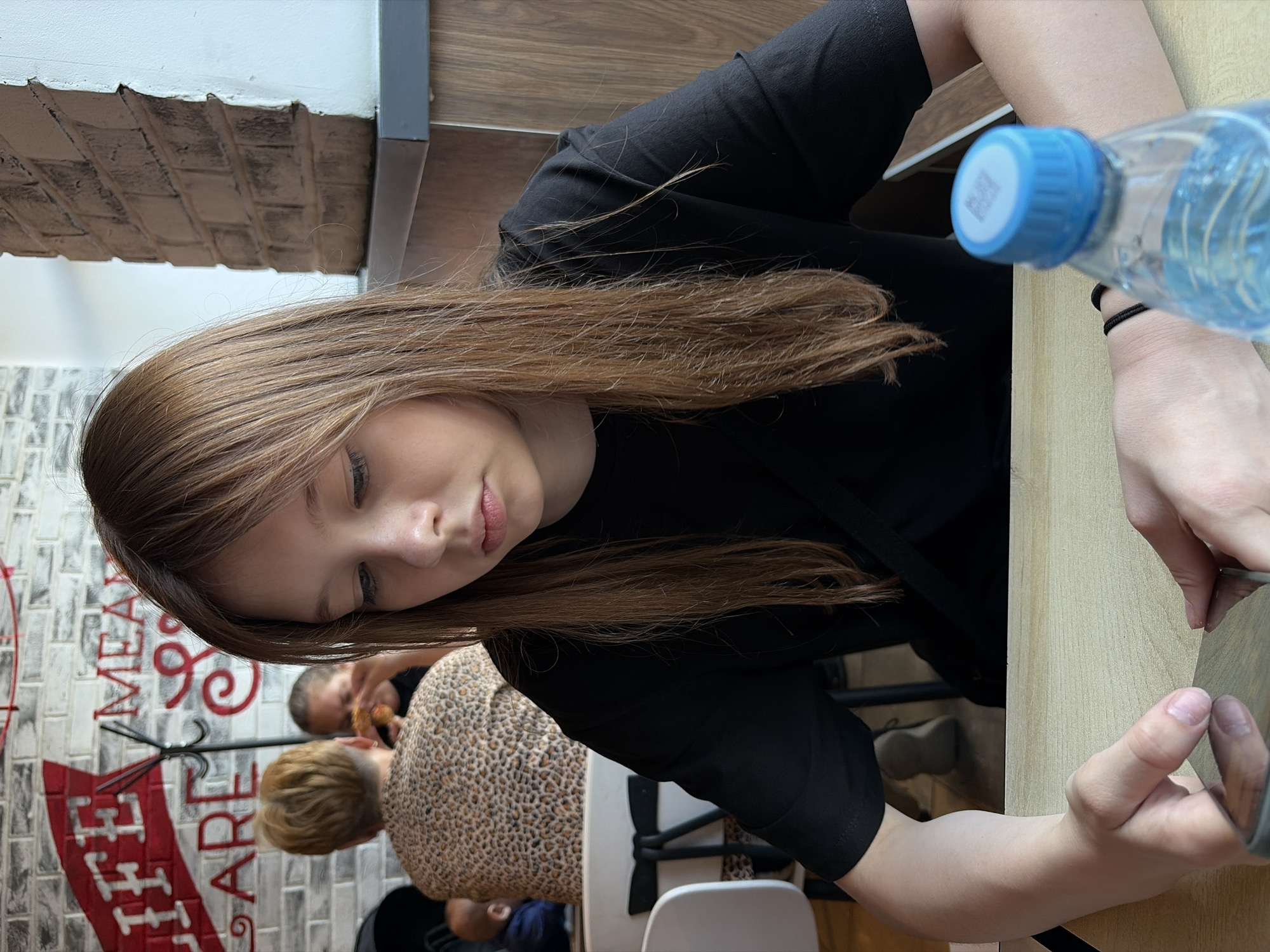 A young Caucasian female with long, light brown hair and bangs is seated at a light-colored wooden table, looking down at a smartphone held in her hands. She is wearing a black t-shirt and a black strap is visible across her chest. To her right, on the table, is a clear plastic water bottle with a blue cap. The background shows a brick wall with white and red painted text, a brick pillar, and other patrons at tables. The overall setting appears to be a casual dining establishment, possibly a fast-food restaurant or cafe, within the city of Chaikovsky, Russia.
