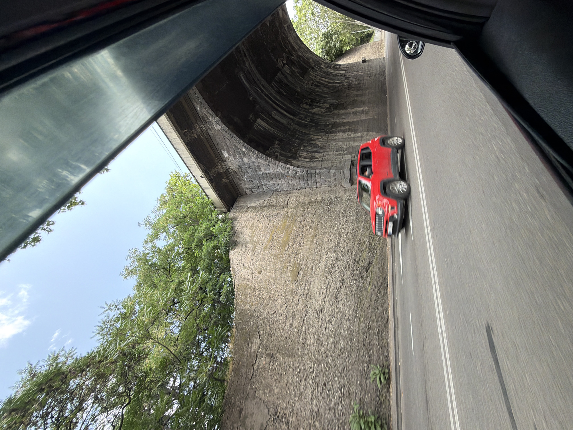 A bright red Jeep Renegade is captured in motion, driving on a road in Tbilisi, Georgia. The vehicle is positioned in the left lane, passing under a large stone arch bridge. The bridge structure is made of rough, grey stone blocks and shows signs of age and wear. To the left of the road, a tall, textured concrete wall rises, supporting dense green trees and foliage. The sky above is a clear, pale blue with wispy white clouds. The photo is taken from inside another vehicle, with the edge of a car window and door visible in the foreground, suggesting the perspective of a passenger. The street has double white lane markers.