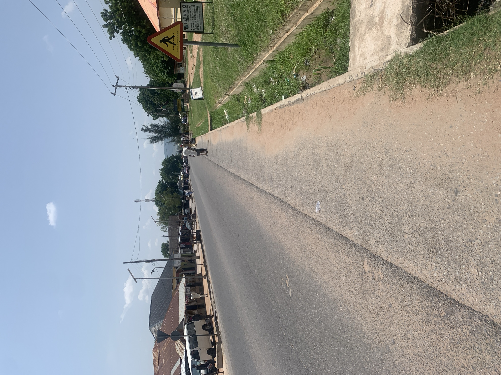 This media file is a photograph of a street scene in Jalingo, Nigeria, taken from a low angle looking down the road. The time of day appears to be daytime, with bright sunlight casting shadows. The weather seems to be clear and fair.
The scene depicts a moderately busy street with several vehicles, including cars and what appear to be vans or minibuses, parked along the side or in motion. There are also a few people visible on the side of the road, some walking and others standing. The buildings lining the street are a mix of structures with corrugated metal roofs and some with more conventional facades. In the foreground, a paved road stretches into the distance, bordered by a sandy shoulder and a grassy verge.
Notable details include a yellow triangular warning sign with a black pictogram of a person walking, indicating a pedestrian crossing. There are also utility poles with numerous wires running overhead, characteristic of many urban or semi-urban areas. One of the buildings has signage that includes the text "BEAUTY MAX SALOON (EVERYTHING BEAUTY)" along with other service descriptions and phone numbers. The overall impression is of a typical day in a bustling commercial or residential area in Nigeria.