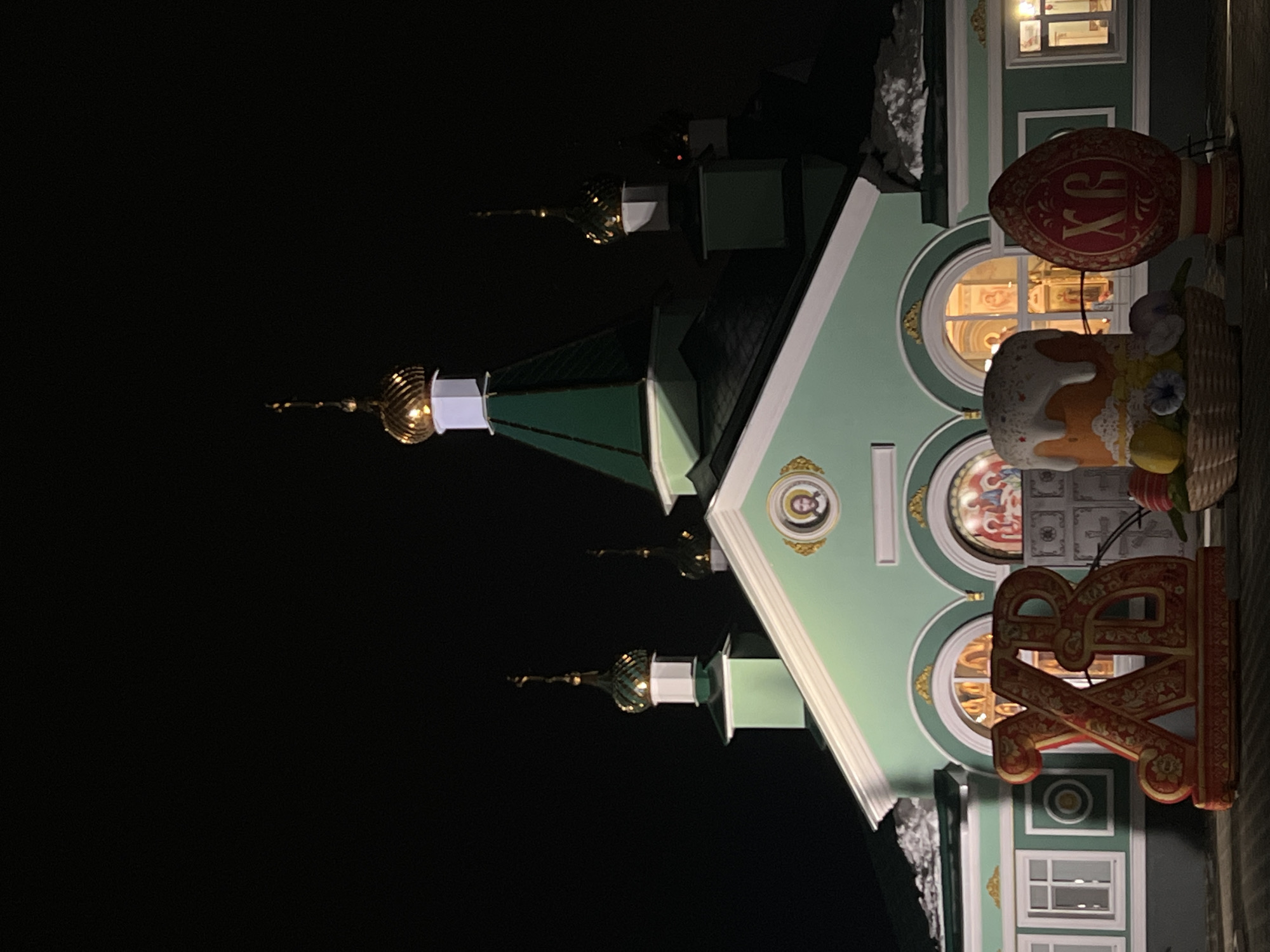A nighttime shot features a green church, likely in Chaikovsky, Russia, with multiple golden domes. Easter-themed decorations, including a painted egg, a basket, and Cyrillic letters, are attached to the side of the church. The surrounding environment is dark.