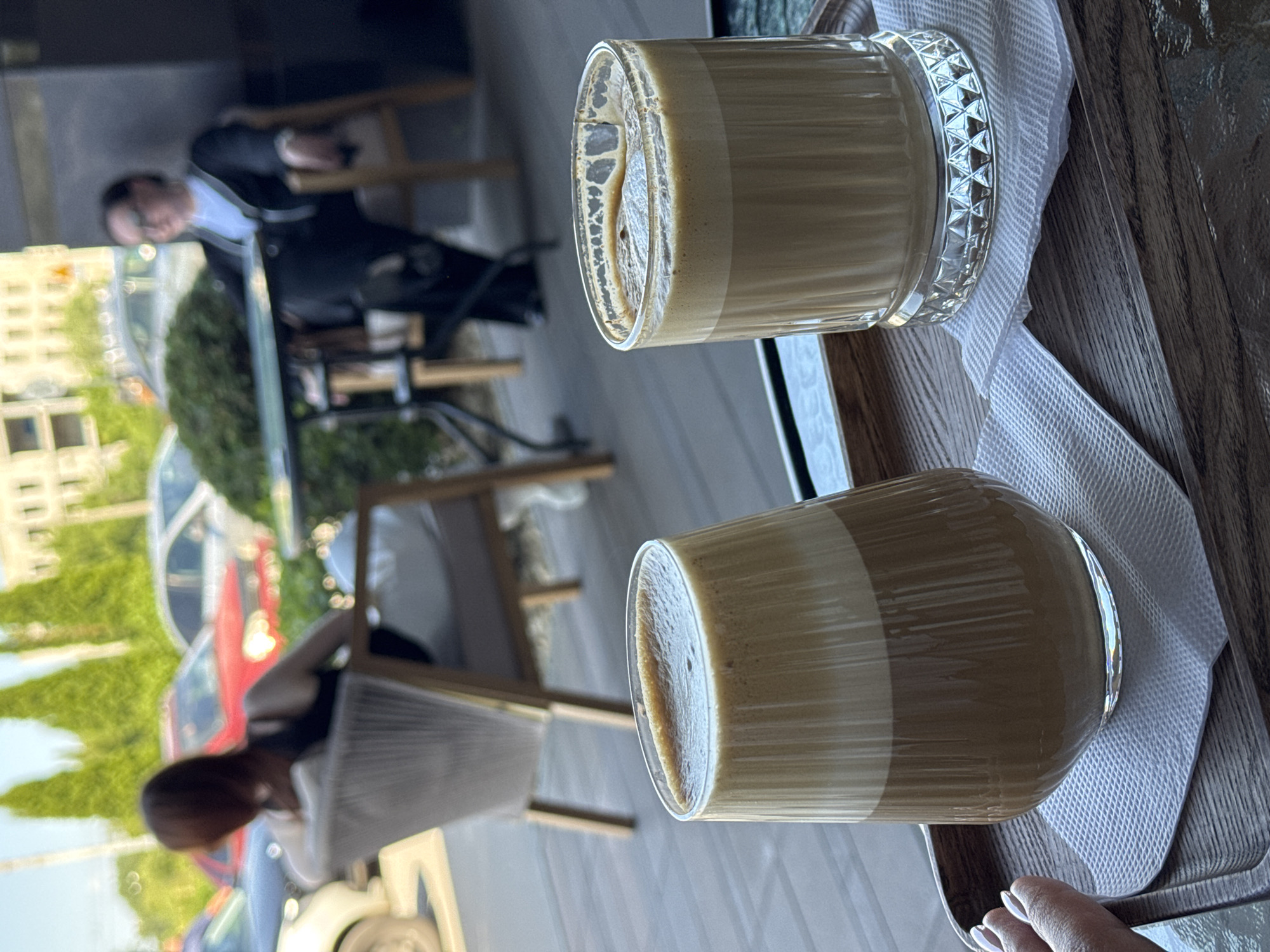Two glasses of iced coffee are displayed on a wooden tray, each placed on a white paper napkin. The glass on the left is wider at the top and tapers towards the bottom, showcasing distinct layers of coffee and foam. The glass on the right is a standard cylindrical shape with decorative ridges around its base. In the background, two individuals are seated at tables outside what appears to be a cafe. One person, viewed from the back, is seated in a white woven chair. The other individual, facing left, is wearing glasses and a dark jacket over a white shirt. Several cars are parked behind them, and a building with multiple windows is visible in the distance, suggesting an urban outdoor setting.