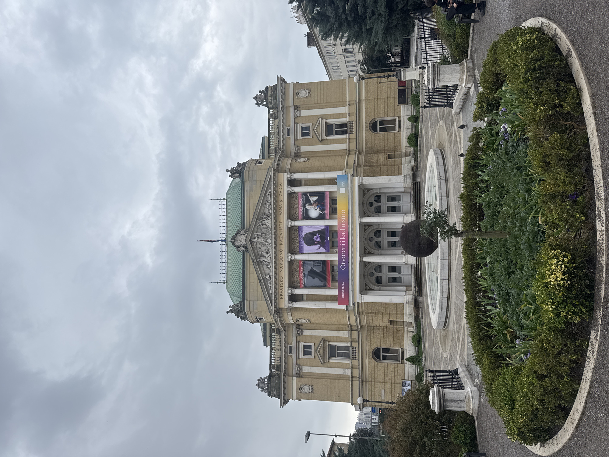 The image shows the Croatian National Theater Ivan pl. Zajc in Rijeka, Croatia. The building is a cream-colored classical structure with a green dome and ornate details. Banners advertising performances, including "Turandot", "Mali Princ", and "Balkanski Spijun" hang from the facade. A fountain and manicured gardens are in front of the building. The sky is overcast.