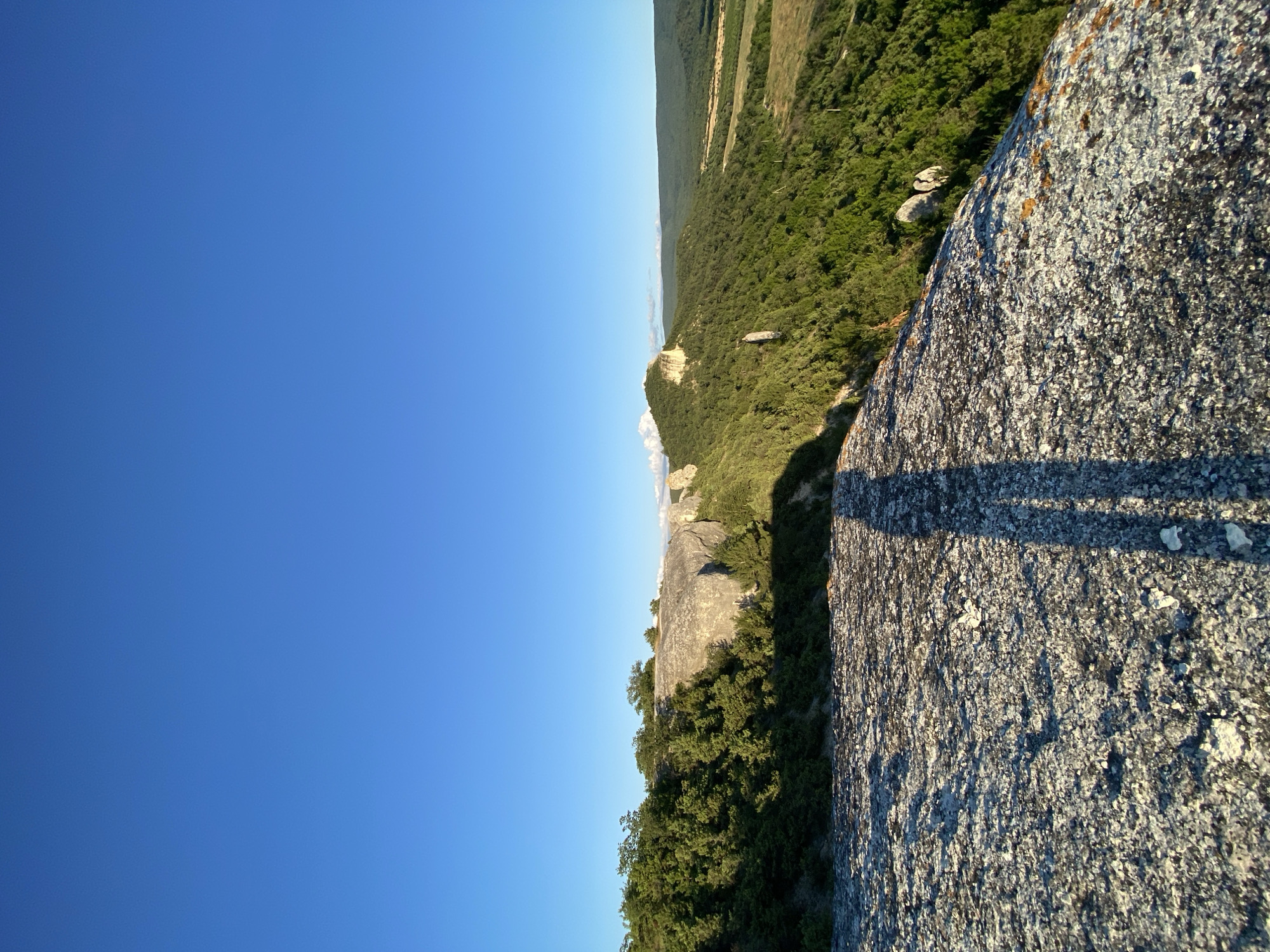 The image captures a scenic landscape with a prominent rock formation in the foreground and a lush green valley stretching into the distance under a clear blue sky. The rock in the foreground, appearing weathered and textured, casts a long shadow, suggesting the presence of a person or object not fully visible in the frame. The shadow's shape indicates it is a human figure, likely standing on the edge of the rock formation, looking out at the vista.
The mid-ground features a dramatic cliffside covered in dense green foliage and punctuated by large, light-colored rocks. These rocks rise above the trees, their surfaces illuminated by sunlight. Further in the distance, rolling hills covered in dark green trees recede into a hazy, verdant landscape. In the far distance, a lighter, cultivated field is visible, contrasting with the natural forest. The sky is a uniform, deep blue, free of clouds, indicative of clear weather.
The scene appears to be a natural park or mountainous region. Given the location provided, this view is likely from the Crimean Mountains near the settlement of Krasnomakskoye, Ukraine. The overall impression is one of natural beauty and a sense of vastness.