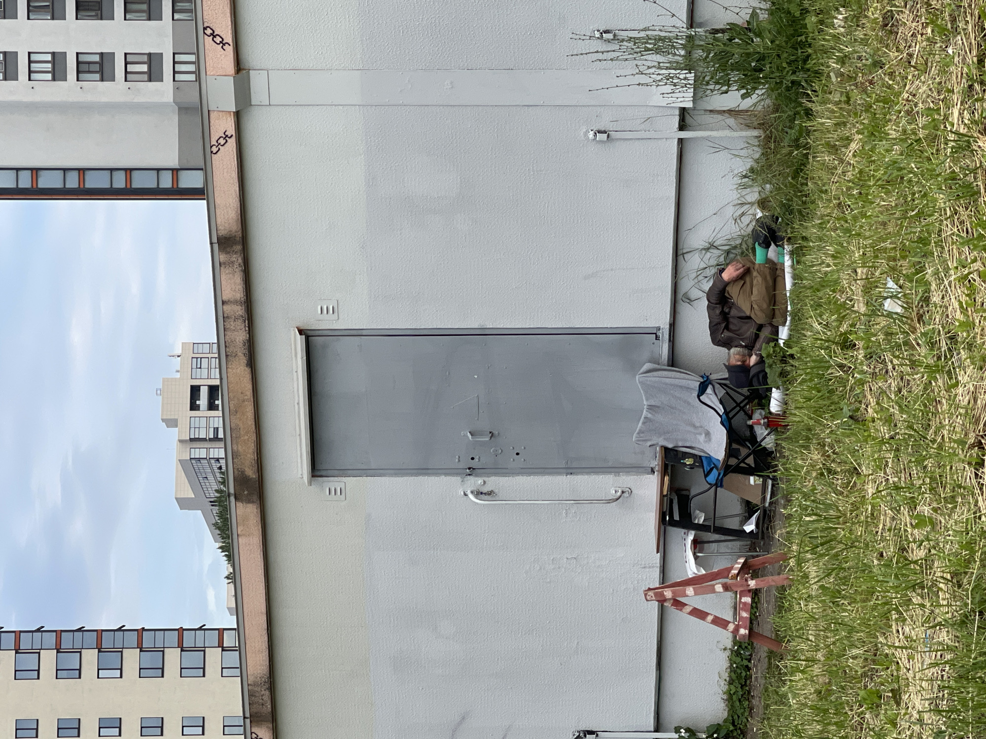 The image shows a person sleeping on the ground in front of a light-colored building with a large gray door. The person is wearing a dark jacket and brown pants, and appears to be lying on a white tarp. To the left of the person, there is a folding chair with a gray towel draped over it, a small table with some items on it, and a red and white striped sawhorse. The background shows parts of tall, modern buildings against a cloudy sky. The area in front of the building is covered in grass.
