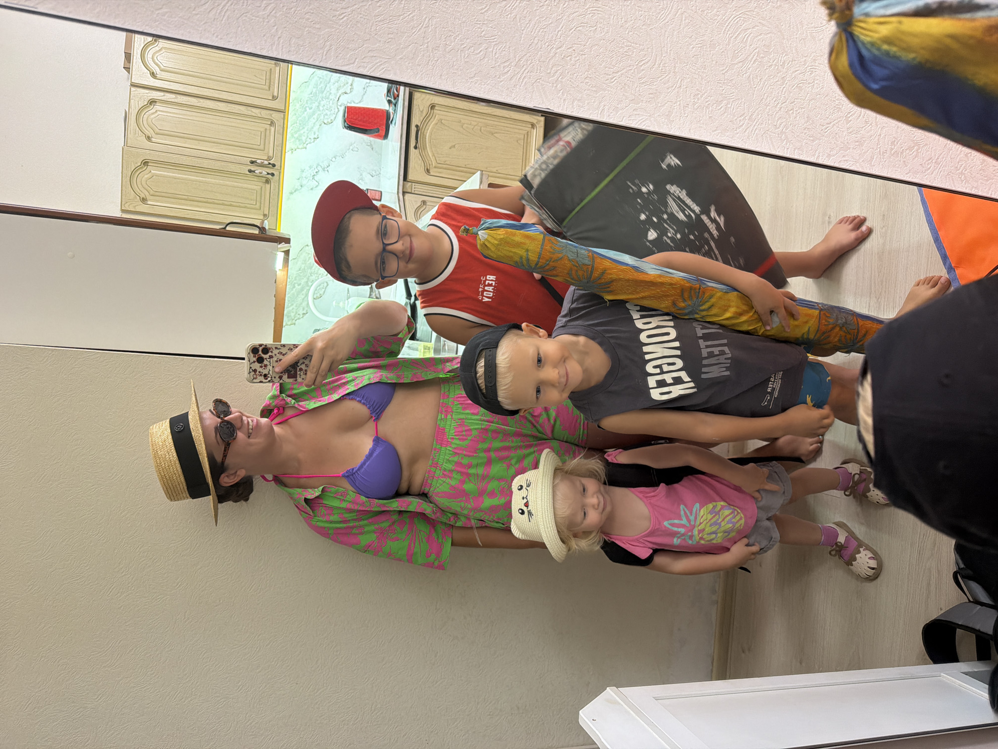 A family of four, including two adults and two children, are posing for a mirror selfie in what appears to be an apartment interior. The woman, on the left, is wearing a purple bikini top and a vibrant pink and green patterned button-down shirt and shorts. She has a straw hat and sunglasses on. In front of her, a young girl in a pink tank top with a pineapple print and gray shorts, wearing a straw hat with a cat design, stands with a backpack. To the right of the woman, a boy in a red basketball-style tank top and a red baseball cap is holding a rolled-up item, possibly a surfboard or yoga mat, and a black case. Next to him, a younger boy in a dark t-shirt and a black baseball cap is also visible. The background shows a kitchen area with light-colored cabinets and countertops, indicating a domestic setting. The floor is light wood. The overall impression is a family preparing for a trip or outing, likely related to a vacation given the attire. The reflection in the mirror includes kitchen cabinets and a sink area. Given the context, this could be an apartment in Sochi, Russia, known for its beaches and outdoor activities.