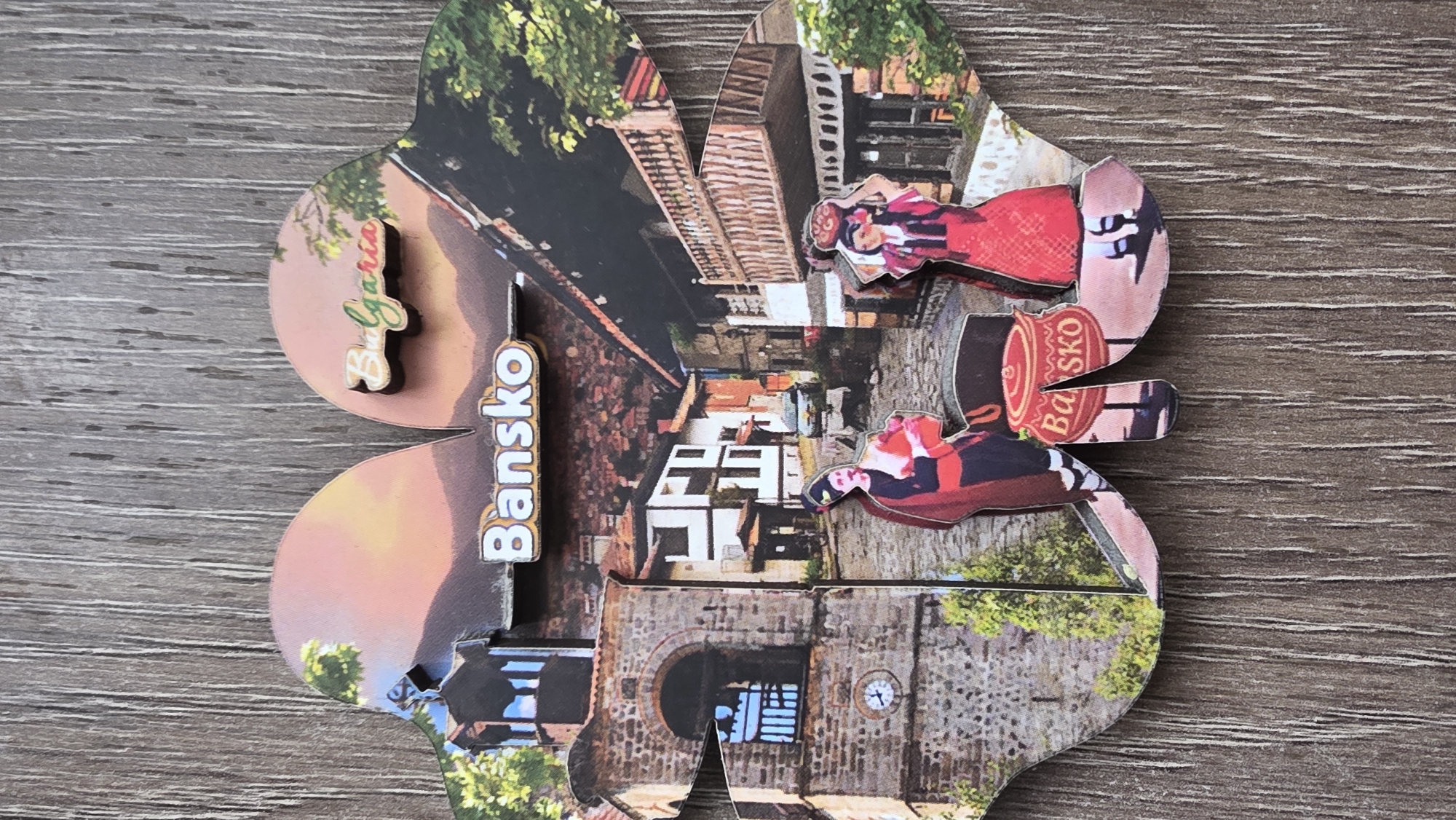 A multi-layered, clover-shaped souvenir, likely a magnet, rests horizontally on a textured wooden surface. The souvenir itself is a collage of images depicting scenes from a town, predominantly featuring traditional European architecture.
The left side of the souvenir prominently displays two raised, wooden text elements. The upper one spells "Bulgaria" in green and orange letters against a background of a soft, orange-pink sky, suggesting a sunset or sunrise. Below it, centered on the souvenir, is the word "Bansko" in white letters with a brown outline.
The images on the souvenir include various buildings, cobblestone streets, and greenery. The bottom left section features a stone clock tower with a small, visible clock. On the right side, two cut-out figures, dressed in traditional folk costumes with red and white elements, are visible. One figure is in the upper right petal, and another is in the lower right, positioned near a red barrel-like object that bears the text "Ba SKO," reinforcing the location. The overall scene captures the essence of a historical town, likely Bansko, Bulgaria, as a travel keepsake.
