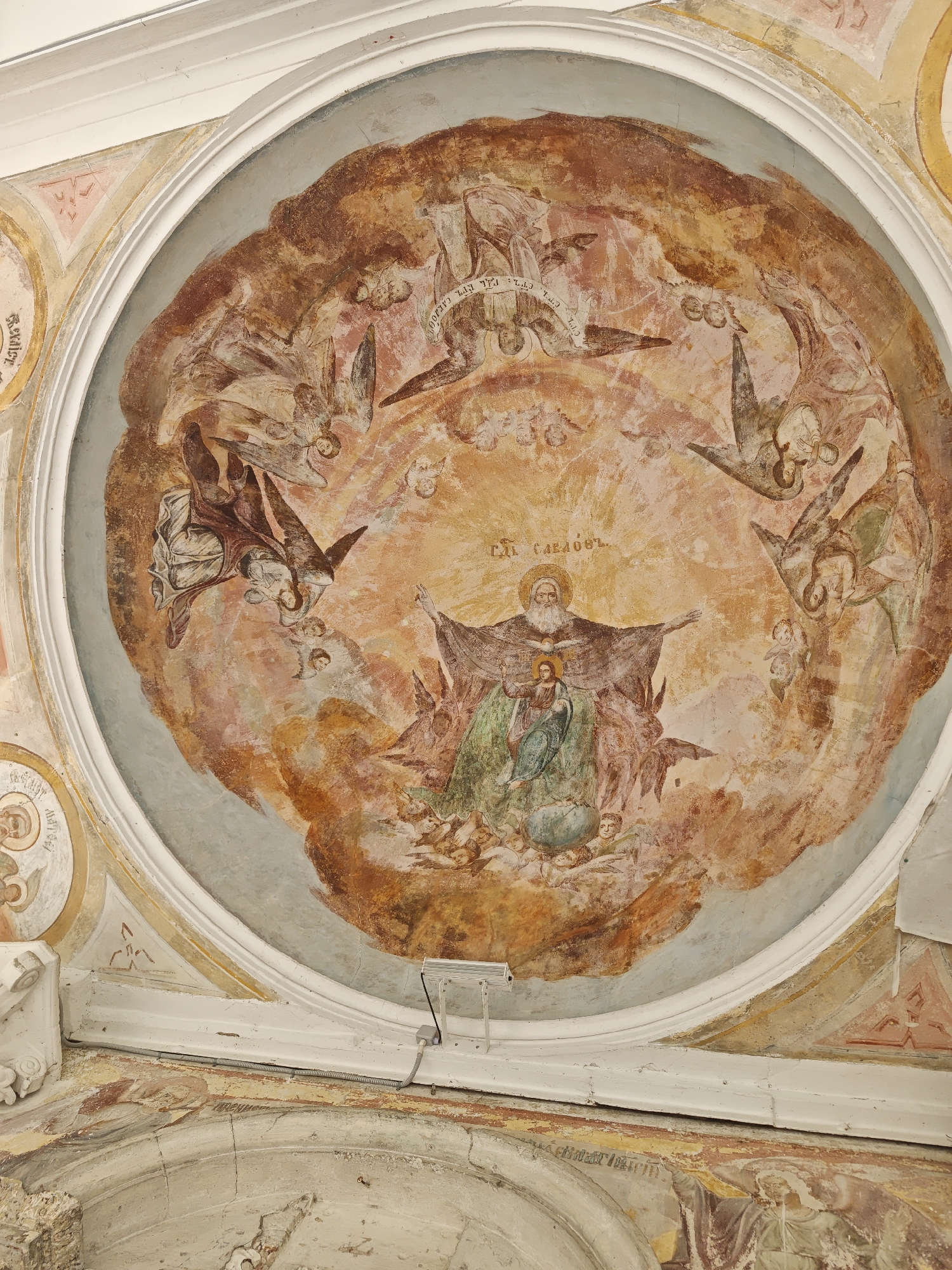 A ceiling fresco depicting a religious scene is located in what appears to be an old building in Selivanovo, Russia. The fresco is enclosed within a circular frame and set against a background of faded, earth-toned colors, with shades of brown, orange, and pink predominating. The central figure is an elderly man with a white beard and halo, holding a younger female figure with a halo. They are standing on a depiction of the earth or a globe, and are surrounded by numerous cherubic figures. Additional figures that appear to be angels with wing details, are depicted around the top half of the circular image. There are inscriptions in what appears to be an ancient, non-English language above the elderly man. The fresco is in an imperfect condition, with some visible signs of aging and degradation. Additionally, there is a modern light fixture mounted underneath the artwork.