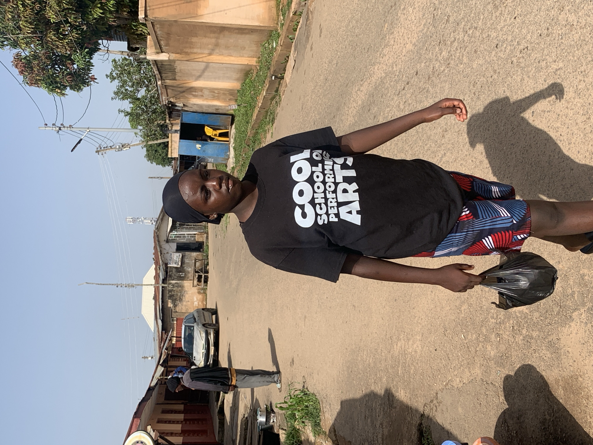 A young Black female walks towards the camera on a dusty, paved road in Jos, Nigeria. She is wearing a black t-shirt with white text that reads "COOL SCHOOL OF PERFORMING ARTS" and patterned shorts. She carries a black plastic bag in her right hand. Behind her, a silver Peugeot 206 car is parked on the side of the road, and several buildings line the street. The architecture of the buildings suggests a mix of residential and commercial properties. The sky is clear and blue, indicating a sunny day. A utility pole with power lines is visible in the background, along with trees and vegetation. A person in a dark jacket and grey pants stands near the car, facing away from the camera. The overall scene depicts a typical day in a Nigerian urban environment.