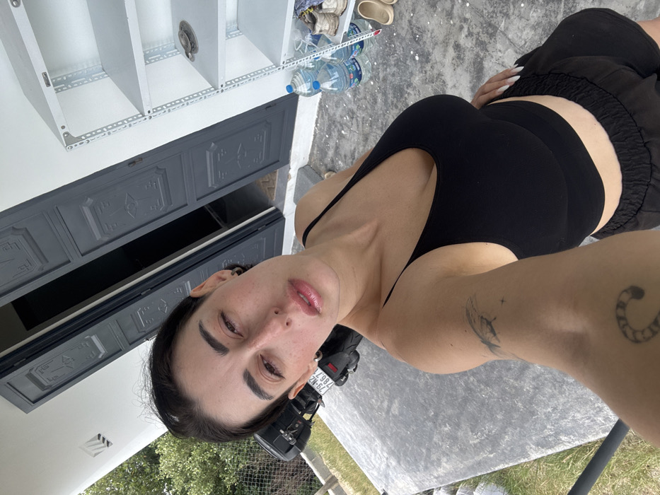 A woman poses in a black top and shorts, showcasing tattoos on her arms, with a neutral expression. The background includes a gray building facade with ornate doors and white shelving units. Clear water bottles sit on the ground. The woman's nails are visible, suggesting meticulous grooming. The scene is outside, with glimpses of greenery and a motorcycle. The image is angled, capturing the environment from a tilted perspective.