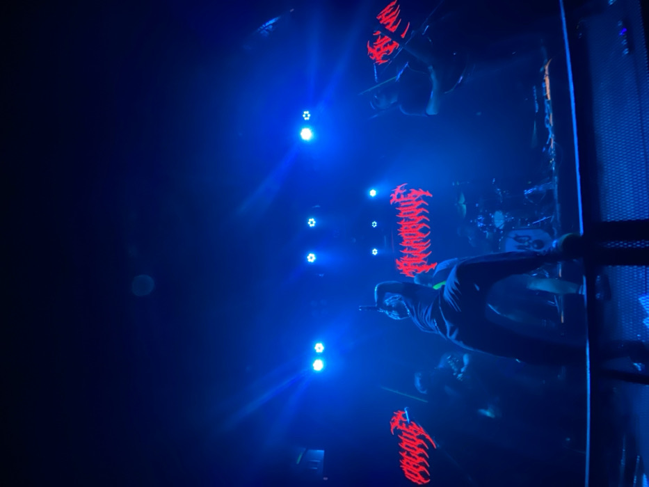 A live musical performance by the band Arcaica is depicted on a stage in a darkened indoor concert venue, likely Uniclub, located in the city of Buenos Aires, Argentina. The scene is primarily illuminated by intense blue stage lighting, with a hazy atmosphere enhancing visible light beams. Three stylized, jagged red logos spelling "ARCAICA" are displayed on the rear wall of the stage, one centrally positioned and two flanking it.
Four band members are present. The central figure, a vocalist, is positioned stage-front, in a dynamic crouch with his right leg extended and left knee bent, holding a microphone in his right hand, facing towards an unseen audience. He is wearing dark attire. To his right, a guitarist, also in dark clothing, plays an electric guitar. To the vocalist's left and slightly behind, a second musician, likely a guitarist or bassist, is partially visible. At the rear-center of the stage, a drummer is seated behind a dark drum kit, with a white kick drum head visible. Multiple overhead blue LED stage lights are directed towards the stage. In the immediate foreground, a dark metal mesh barrier separates the audience perspective from the stage. The band members are actively performing their respective roles.
