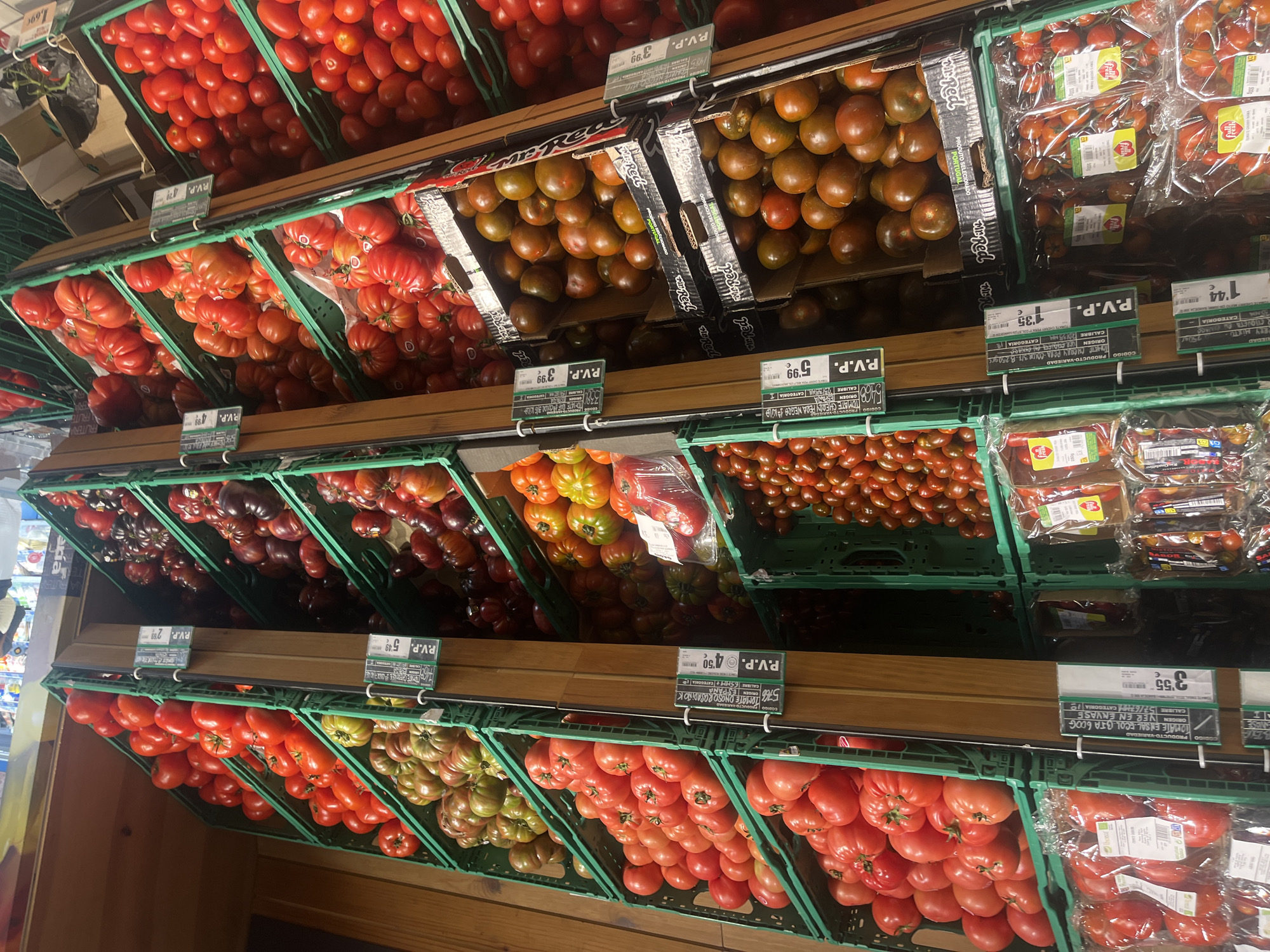 The image shows a supermarket produce aisle displaying a variety of tomatoes. The tomatoes are arranged in green plastic crates on wooden shelves. The varieties include large red tomatoes, some with ribbed surfaces, smaller red cherry tomatoes, and darker, multi-colored heirloom tomatoes. Price labels are visible above some of the crates, with prices indicated in Euros. One label mentions "P.V.P." which stands for "Precio de Venta al Público" (Retail Sale Price). The supermarket appears to be well-lit, and the overall impression is one of a clean and organized retail environment. There are no people visible in the immediate foreground, but the presence of multiple shelves filled with produce suggests a typical retail grocery store. The specific location in A Coruña, Spain, cannot be determined solely from the visual information in the image.