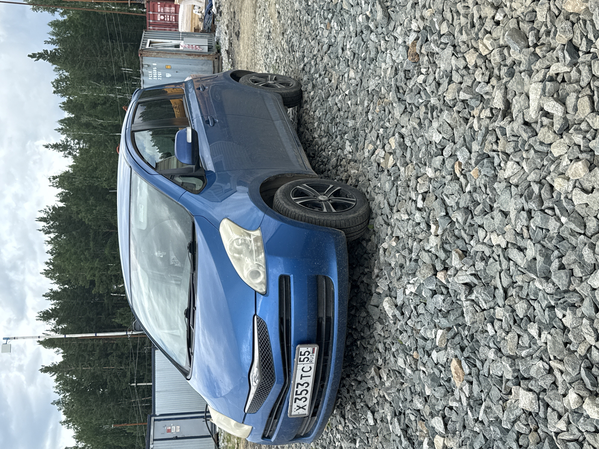 The image shows a blue Toyota Ractis parked on a gravel surface in what appears to be an industrial or construction area. In the background, there are shipping containers, some with visible markings such as "PCLC U 205805 0 2261" and "162276". Behind the containers, a dense forest of pine trees is visible under a cloudy sky. A tall, slender structure with a white antenna on top stands to the left of the car. The car's license plate reads "X353TC 55" with "RUS" beneath it. The scene suggests a location in Leninsk, Russia, possibly a port or a storage facility given the presence of containers and industrial elements.