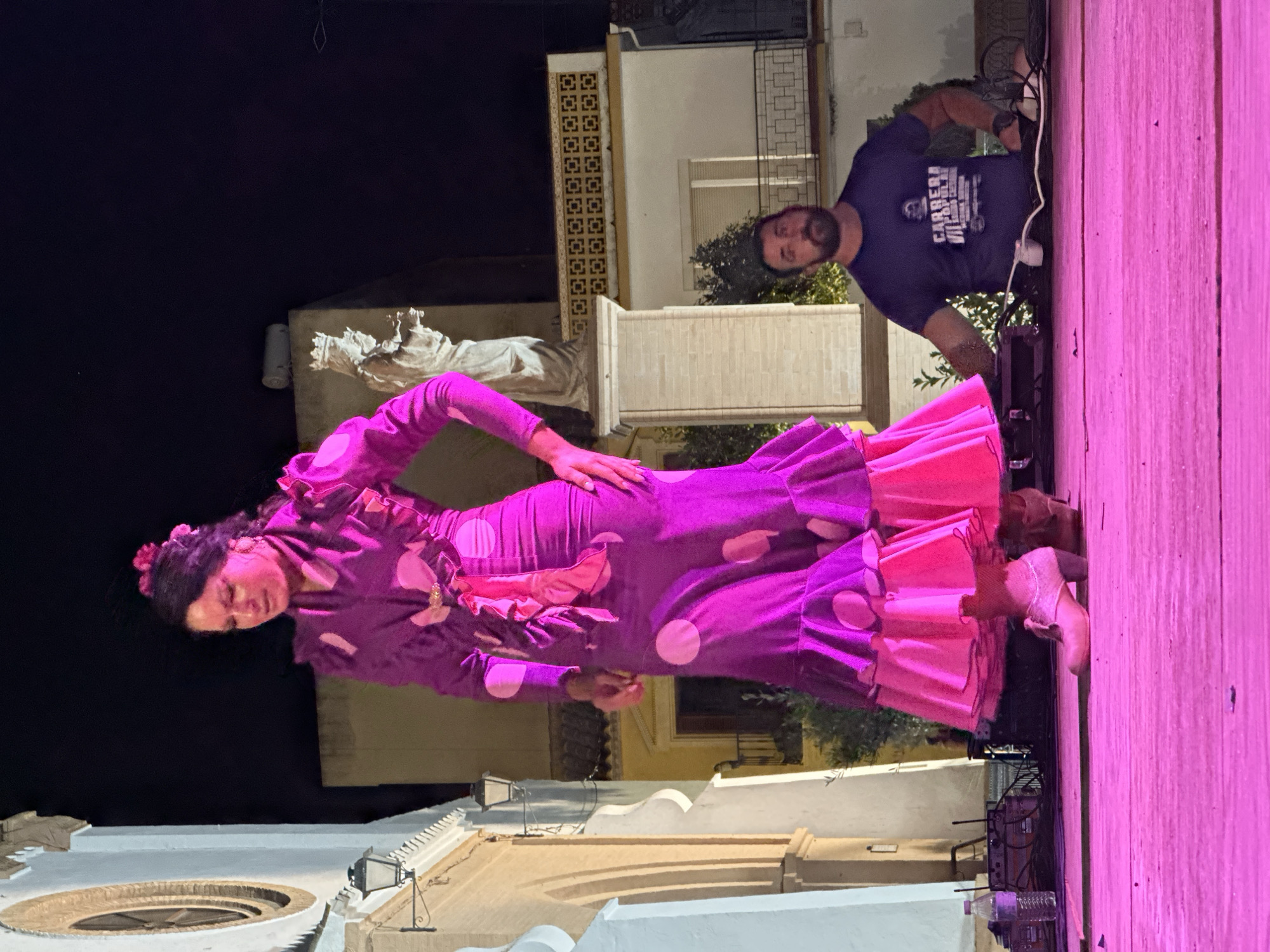 The image captures a nighttime outdoor performance in San José del Valle, Spain. A woman dressed in a vibrant purple and pink polka-dotted flamenco dress with ruffled layers stands center-stage, her gaze directed downwards in concentration. Her hair is styled up and adorned with pink flowers, and she wears large hoop earrings. She is barefooted, but wearing flamenco shoes. To the right of the frame, a man with a beard wearing a dark blue t-shirt with text and graphics is seated behind a DJ setup, looking towards the performer. He is positioned lower on the stage, with his arms resting on the equipment. The stage itself is a wooden platform bathed in bright pink light. In the background, a white building with arched doorways and decorative lanterns is visible, along with a statue of two figures wearing crowns. Further back, another building with a balcony and a patterned frieze can be seen. The overall scene suggests a cultural event or festival taking place in the town.