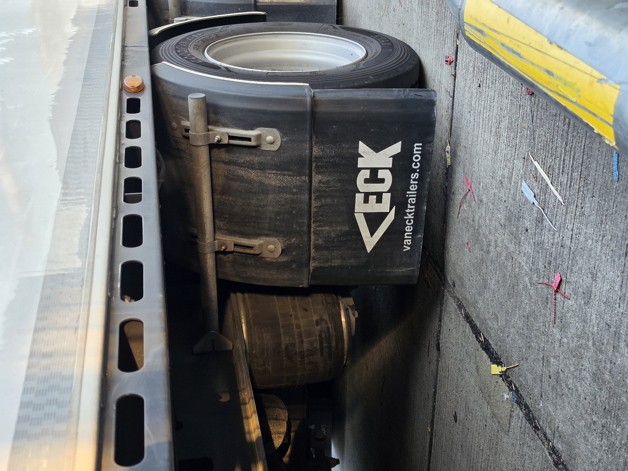 The image shows the underside of a truck trailer, specifically the rear wheel and mudguard. The mudguard is black with white lettering that reads "VECK" and "vanecktrolers.com". There are also some small red and yellow plastic tags scattered on the concrete ground. The trailer itself is silver and reflective. The lighting suggests it is daytime. The image was taken in Frankfurt, Germany.