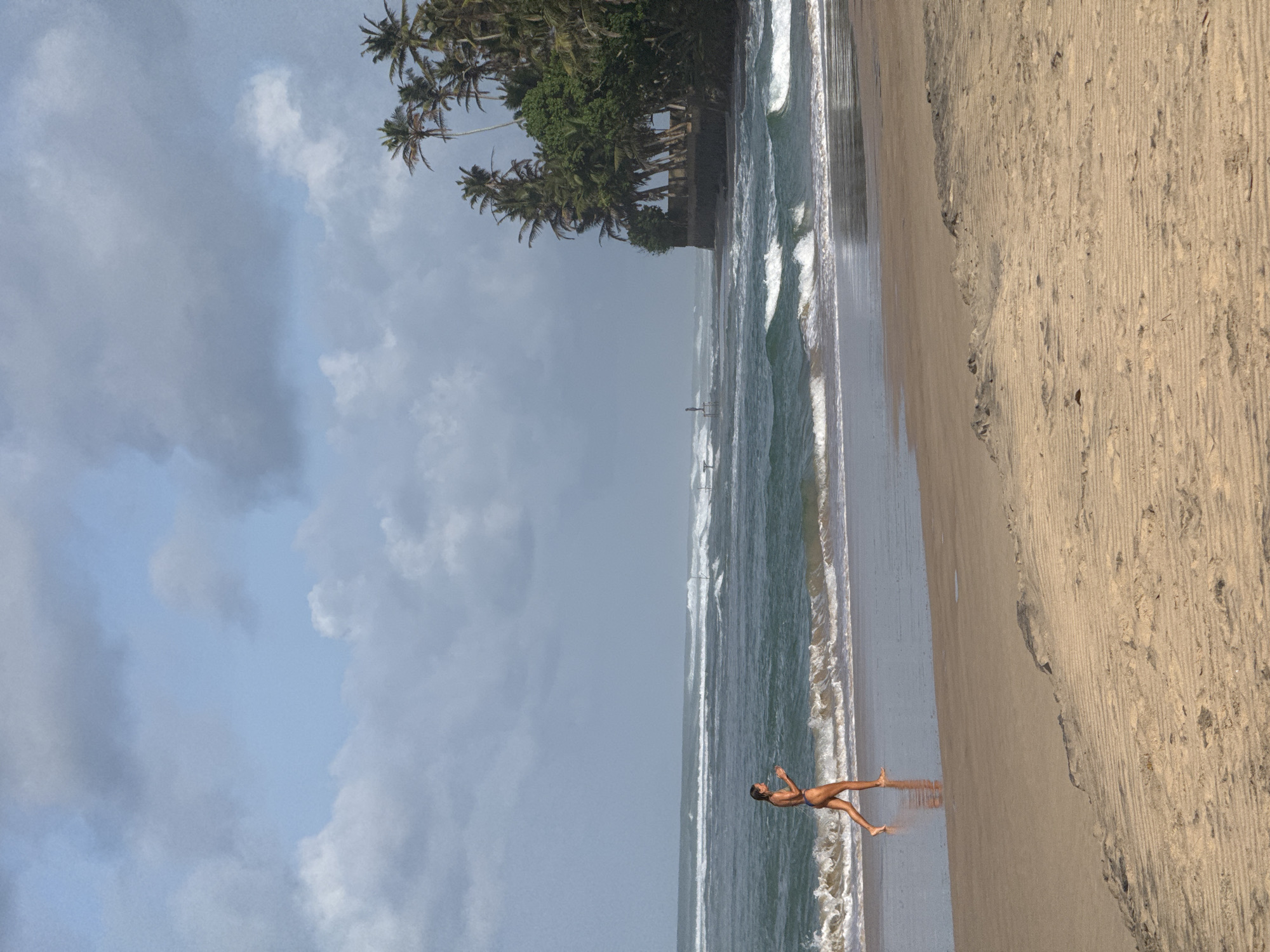 A person is jogging on a sandy beach in Kathaluwa, Sri Lanka. The person is wearing a dark bikini and is running along the waterline, with their reflection visible in the wet sand. The ocean is a deep blue-green, with white waves breaking on the shore. In the background, there are palm trees and lush vegetation lining the coast, with a cloudy sky overhead. The beach is wide and sandy, with the sand showing patterns from the receding tide. The overall scene conveys a sense of a serene and active coastal environment.