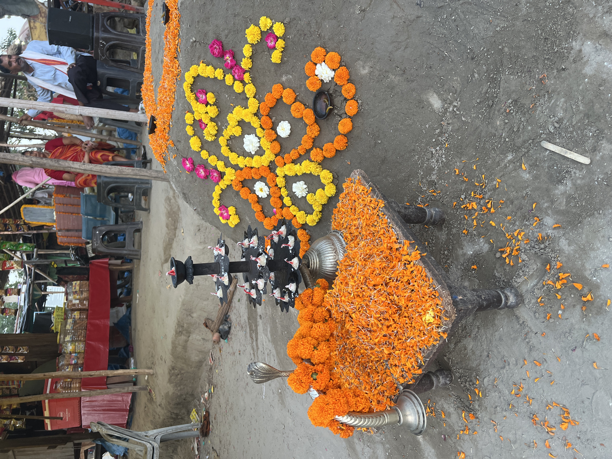 The image depicts a scene in Vrindavan, India. In the foreground, there is an altar-like structure adorned with marigold flowers. The flowers are also arranged in garlands and patterns on the ground surrounding the structure. A multi-tiered oil lamp stand is also present.
In the background, there are people seated on chairs. One man is wearing a white shirt and what appears to be a shawl or sash. There are vendor booths and other commercial structures also present in the background.
The ground is paved, suggesting an urban or semi-urban setting. The overall impression is one of a religious or ceremonial event taking place in a public space.