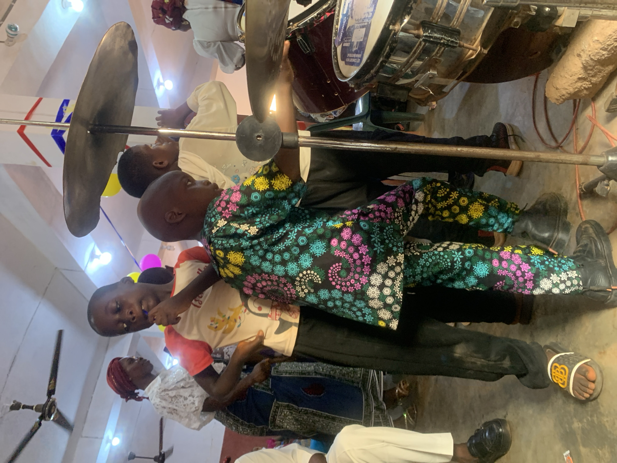 The image depicts a group of people gathered in what appears to be a musical setting in Nsugpe, Nigeria. Several individuals are focused on playing musical instruments, including a cymbal and drums. One child is wearing a shirt with colorful snowflake patterns. Other adults are present, some seemingly observing or assisting. The environment appears to be indoors, possibly a religious or community hall, judging by the decorations, including a flag and balloons. There is a focus on music performance.