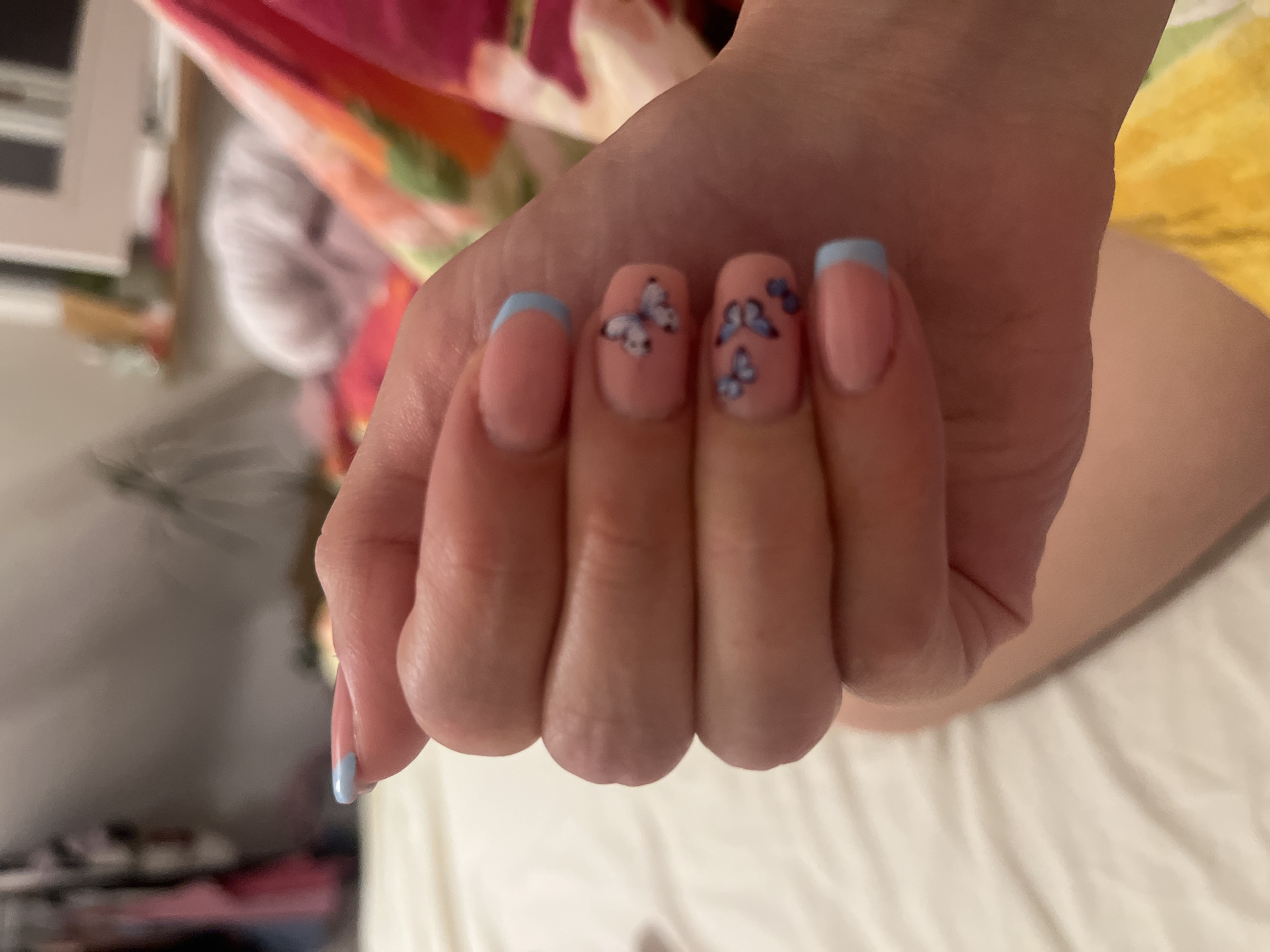 A hand with manicured nails is visible. The nails are painted in a pale pink color with a light blue French tip on the thumb, index, and pinky fingers. The middle and ring fingers are adorned with butterfly nail art. The hand is positioned against a blurred background of what appears to be bedding and a bedside table. The location of Saarbrücken, Germany is unknown.