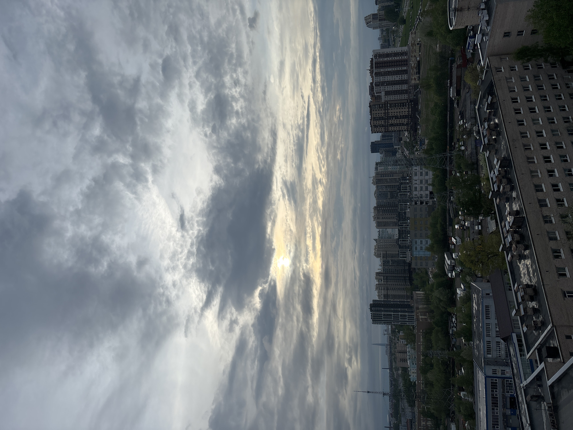 The image depicts a cityscape under a cloudy sky. A multitude of buildings of varying heights and architectural styles form the urban landscape. A transmission tower stands prominently in the distance. The sun, obscured by clouds, casts light across the scene. A mixture of older brick buildings and more modern high-rise structures is visible. The city appears to be Unknown, Unknown.