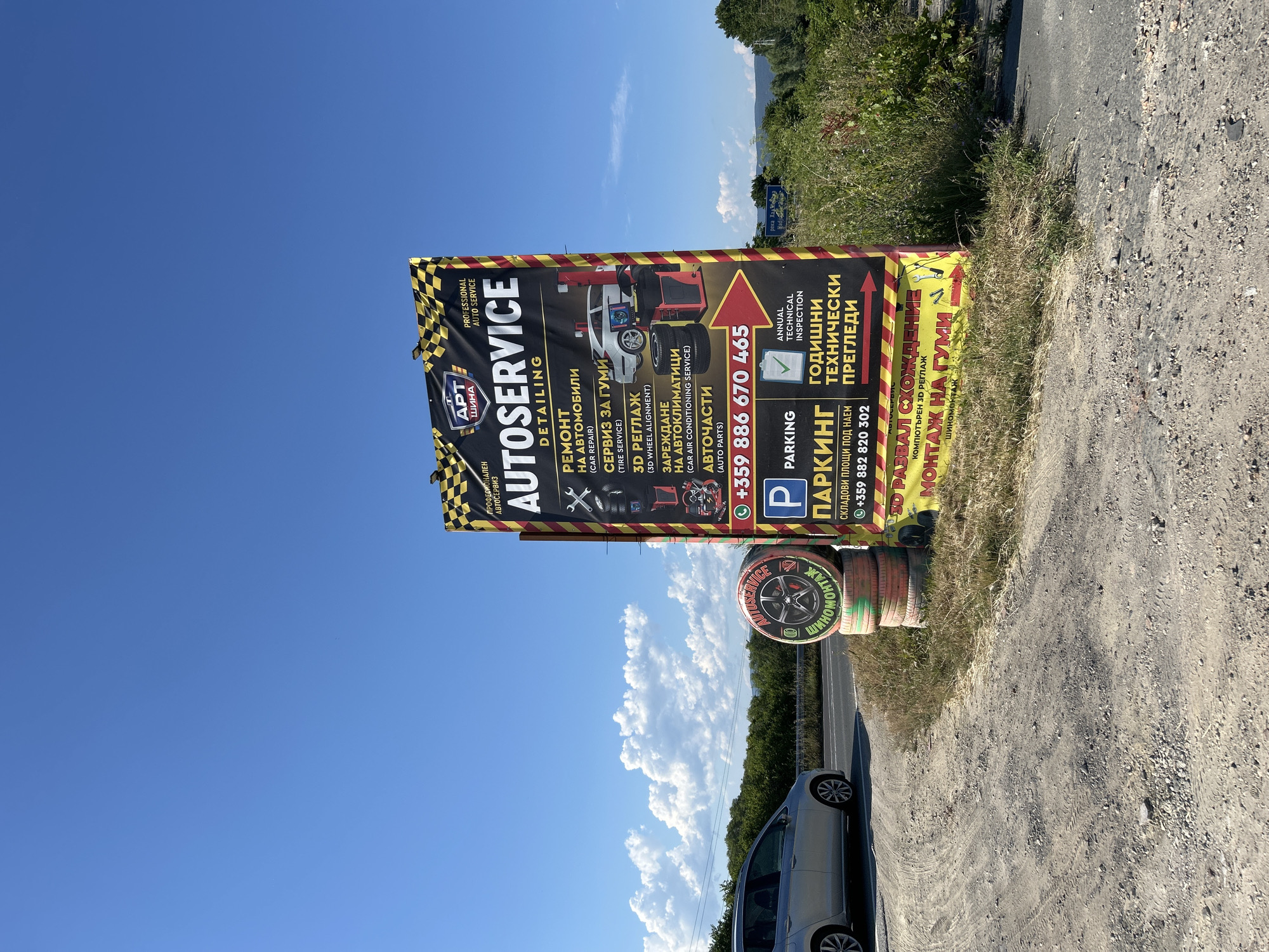 This is a medium-angle, eye-level shot of a large, black and yellow checkered billboard advertising an auto service business, located on the side of a road. The billboard, which has the text "AUTOSERVICE DETAILING" prominently displayed, lists various services offered, including car repair, tire service, 3D wheel alignment, air conditioning service, auto parts, parking, and annual technical inspection. The phone number "+359 886 670 465" is also visible. Below this, there is text for "РАЗВАЛ СХОЖДЕНИЕ" and "МОНТАЖ НА ГУМИ", which are Bulgarian for "wheel alignment" and "tire fitting". The billboard is situated in a rural or semi-rural area with a clear blue sky and white clouds. In the foreground, to the left, is a silver hatchback car parked on a dirt and gravel area next to the road. Several used tires are stacked behind the billboard. In the background, there is green vegetation and trees, with hills or mountains visible in the distance. A small blue road sign in the far distance appears to indicate a river. The location is identified as being in Nesebar, Bulgaria.