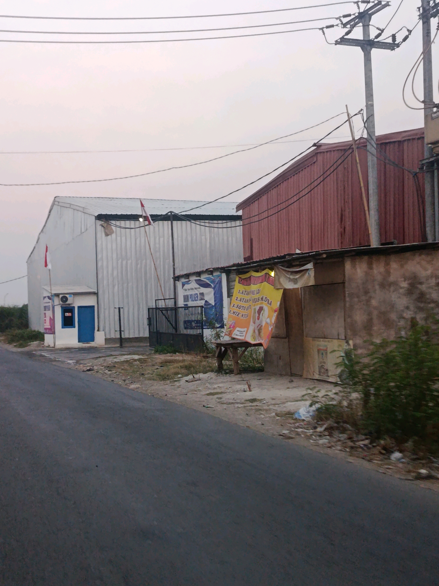 The image depicts an outdoor scene in Bunibakti, Indonesia, with a focus on a street adjacent to industrial or commercial buildings. In the foreground, a paved road curves gently to the left. To the right of the road, a small, dilapidated building with a brown, weathered facade stands next to a larger, corrugated red metal structure. A yellow banner advertising food, with text in Indonesian and images of food items and people, hangs from the front of the small building. Various debris and vegetation are scattered around the base of these structures.
Further to the left, a tall, white corrugated metal building with a sloped roof dominates the background. In front of this building, a small, white security booth with a blue door and an air conditioning unit is visible. Two Indonesian flags are flown from poles attached to the white building, one on the security booth and another on a taller pole. A dark metal fence with a gate separates the security booth area from the red metal building. Overhead, power lines crisscross the sky, supported by a concrete utility pole. The sky is overcast with a pale, diffused light, suggesting it might be early morning or late afternoon. No people are clearly visible in the image, but there are signs of human activity through the presence of the buildings, flags, banner, and road.