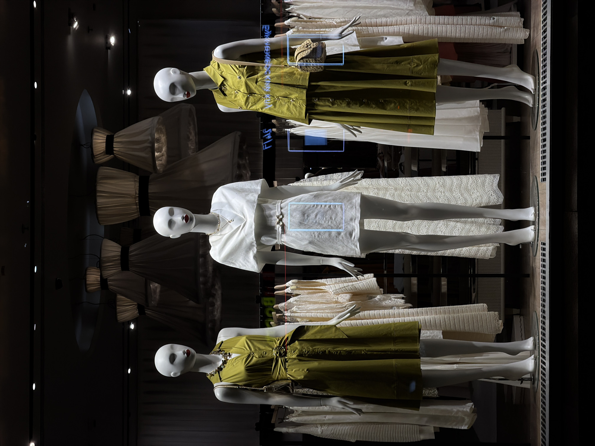 A shop window display in Moscow, Russia, features three full-body, white female mannequins with red painted lips, showcasing fashion attire against a dark retail interior. The mannequin on the left wears a sleeveless, olive-green midi dress with a collared V-neck, button-front closure, and a wide matching belt secured by a large circular buckle. She accessorizes with a multi-strand beaded necklace in earthy tones and a small, olive-green handbag. The central mannequin presents a white, short-sleeved top with a draped V-neckline, paired with a short, white textured skirt cinched with a thin, gold-toned belt featuring a bow-like buckle. She wears a delicate gold-toned necklace. The mannequin on the right is dressed in a two-piece olive-green ensemble, consisting of a sleeveless collared shirt with chest pockets and a matching midi skirt with button detailing. She carries a small, woven straw-colored handbag and wears a delicate gold-toned necklace.
Behind the mannequins, racks display numerous light-colored garments, predominantly white, cream, and beige, including dresses and tops. The ceiling above features several large, inverted bell-shaped decorative lamps with pleated fabric shades and dark banding, alongside recessed spotlights. The floor in the immediate foreground includes a metal grate. Faint, light blue semi-transparent rectangular overlays with indistinct text and what appear to be reflections of "Flas" and "VIBE ARO" are visible across the glass surface of the display.