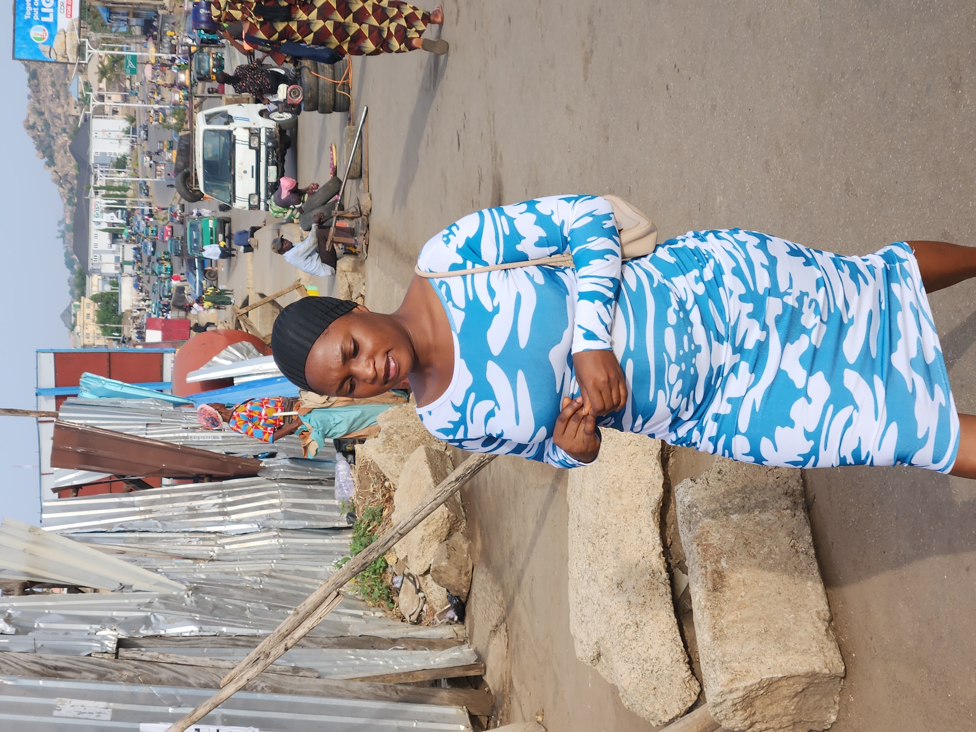 A medium shot depicts a woman wearing a blue and white patterned dress and a black head covering, standing on a street in what appears to be Jos, Nigeria. She has a purse slung over her shoulder. Behind her, there is a scene of urban activity with several cars, a truck, and other vehicles visible. In the background, a hill rises in the distance. To the left of the woman, there is a makeshift structure constructed from corrugated metal. The ground appears to be uneven and dusty. Several people are visible in the background, adding to the bustling street scene.