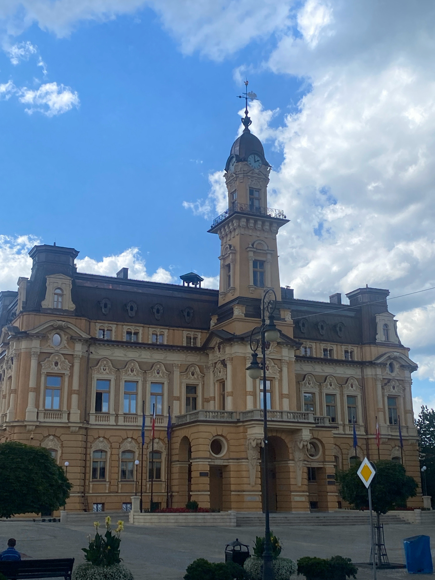 The image shows the facade of the Nowy Sącz Town Hall, a historic building located in the city of Nowy Sącz, Poland. The building features a prominent clock tower with a weather vane at the top, intricate architectural details, and a pale yellow facade. The sky above is a vibrant blue with scattered white clouds. In the foreground, there is a paved plaza with several flags flying on poles. A lamppost stands in front of the building, and to the left, a person sits on a bench. Greenery, including trees and flowerbeds, is visible around the plaza. The overall scene depicts a clear day in a public square in front of a significant historical landmark.