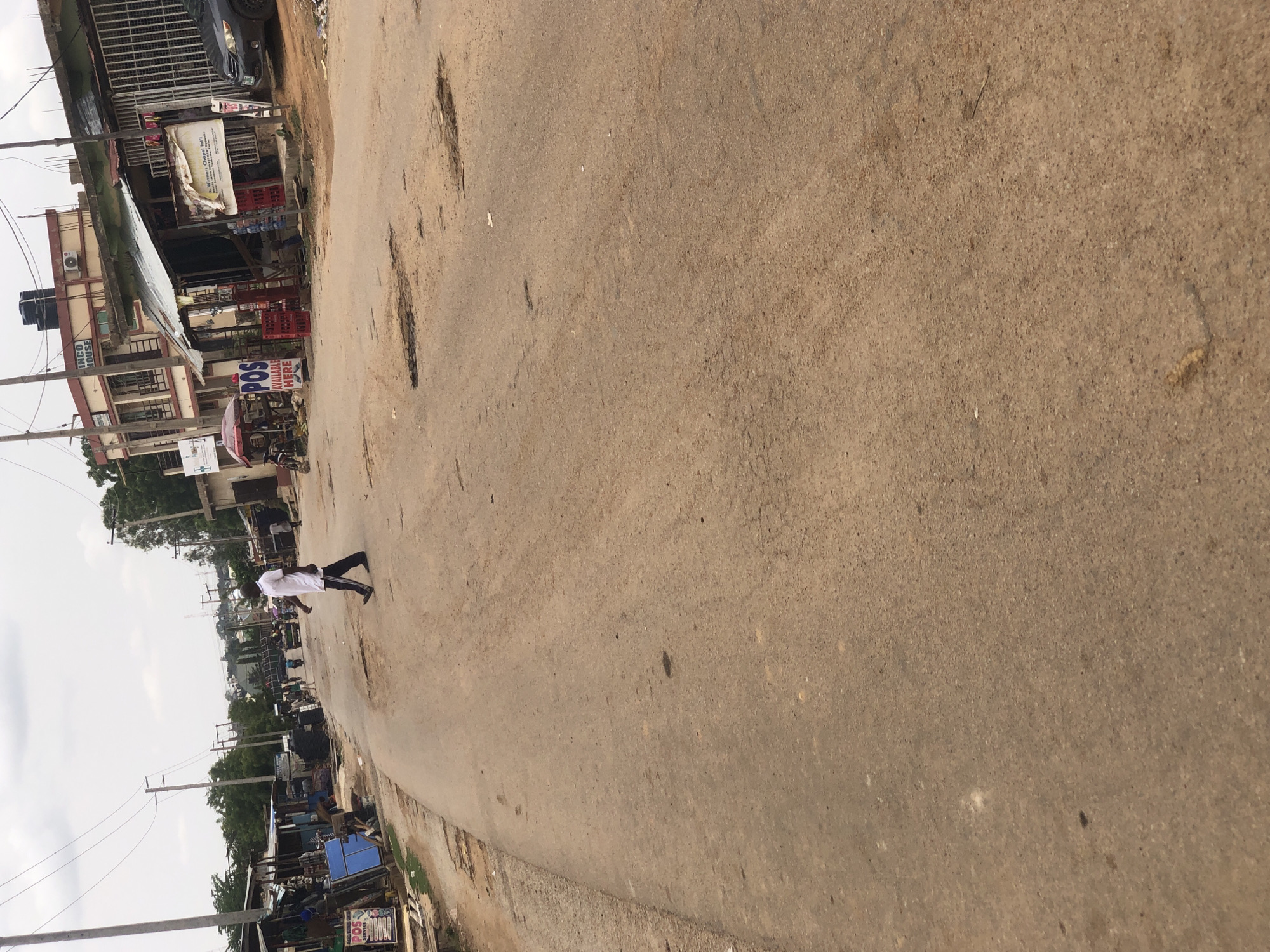 A street scene in Hanwa, Nigeria. The paved road, heavily pitted with potholes, dominates the frame. A single person is walking on the right side of the road, away from the camera. The person is wearing a white top and dark pants. The roadside contains a mix of one and two-story buildings. There are commercial buildings/shops visible. Powerlines and utility poles are also visible. The sky is overcast.