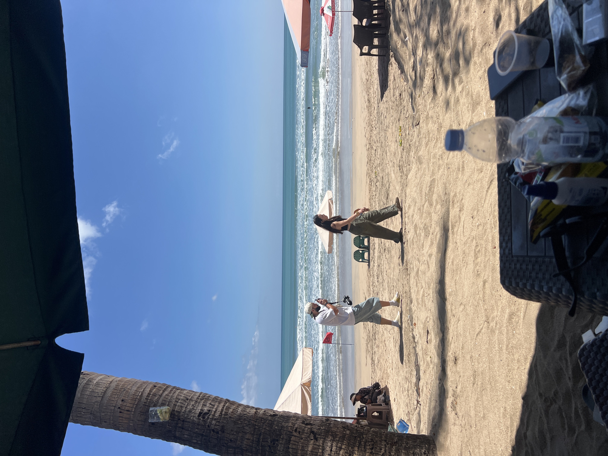 In Kuta, Indonesia, a bright, sunny day unfolds on a sandy beach, with the turquoise ocean stretching to the horizon under a clear blue sky dotted with white clouds. A large palm tree, a prominent feature on the left, casts a shadow, partially obscuring a dark green beach umbrella overhead.
On the sandy shore, two individuals are captured mid-stride. A man, wearing a white t-shirt, light blue pants, and a cap, holds a camera, appearing to take a photo. He is walking from left to right. Beside him, a woman in a dark top and loose olive green pants walks in the same direction, her gaze directed forward.
Further down the beach, beach chairs and umbrellas suggest a relaxed atmosphere. Two empty plastic chairs are arranged near a light-colored umbrella. To the right, a larger umbrella, predominantly orange and white, shades a cluster of dark chairs.
In the foreground, a dark, woven table is set up, strewn with various items. A partially filled plastic water bottle with a blue cap stands prominently, accompanied by a clear plastic cup, a smartphone, and other unidentifiable objects wrapped in plastic. The sand is well-trodden, indicating a popular area. A small red flag planted in the sand behind the man with the camera marks a point on the beach. The ocean is moderately active, with gentle waves rolling towards the shore. A solitary figure can be seen in the water.