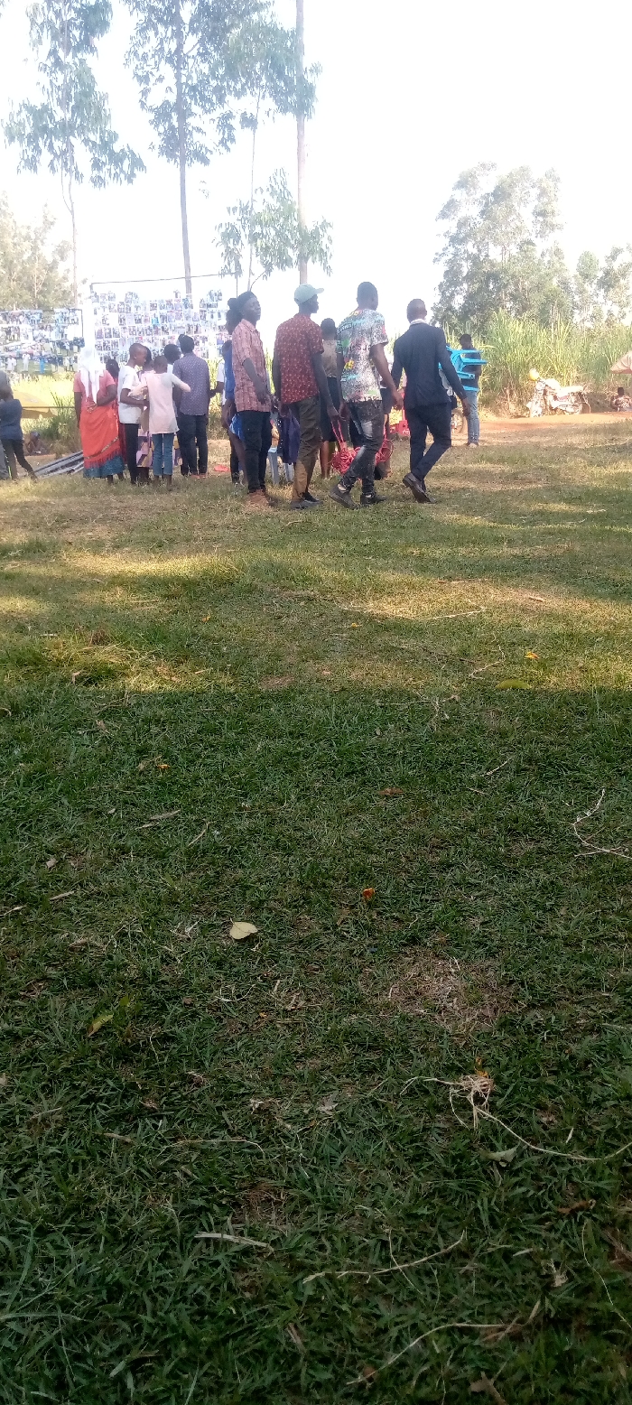 This media file captures an outdoor community gathering in Burundu, Kenya, under bright, clear daylight conditions.
**Subjects**: The primary subjects are a group of approximately 15-20 individuals, mostly adults, with at least one child visible, standing on a grassy field. They are predominantly seen from behind or the side, looking towards a large display board on the left side of the frame. Clothing appears casual for most, with one individual dressed in a dark suit. One woman wears a red dress and a white head covering.
**Objects**: A prominent white board or structure, covered with numerous small photographs arranged in a grid, serves as a focal point for the gathering. The foreground is dominated by lush green grass with some scattered debris. In the background, tall, slender trees with sparse foliage stand against a very bright sky. On the far right, dense green vegetation and what appears to be a dirt path or bare ground are visible, along with a glimpse of a motorbike.
**Scene and Setting**: The scene is set in a sunny, open grassy area, possibly a field or a compound, in Burundu, Kenya. The bright sky suggests a clear day, possibly mid-morning or afternoon, given the shadows on the grass. The tall trees suggest a rural or semi-rural environment.
**Activity or Event**: The people are gathered around and seemingly viewing the display board covered with photographs. This suggests a community event, possibly a memorial, a school function, a local exhibition, or a celebration where images are being showcased or remembered. The general atmosphere appears calm and observant.
**Notable Details**: The intense brightness of the sky indicates strong sunlight. The display board with multiple small pictures is a significant detail, hinting at the purpose of the gathering. There is no clearly visible text on the board or elsewhere that can be deciphered from this distance.