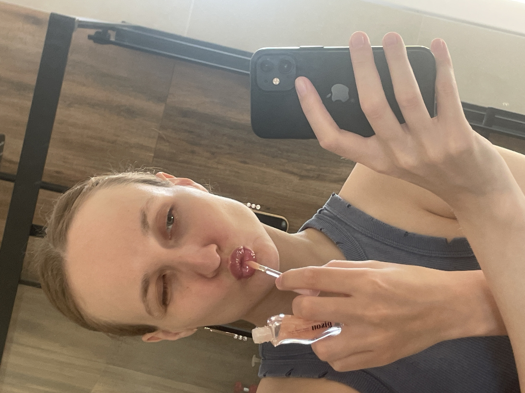 A light-skinned woman is taking a selfie while applying lip gloss. She is holding a black iPhone in her left hand and the lip gloss applicator in her right. Her lips are shiny with lip gloss, and she is wearing a gray tank top. The background appears to be a room with wood-paneled walls and a metal rack or bar. There is no visual evidence to suggest the location is Подшивалово, Russia.