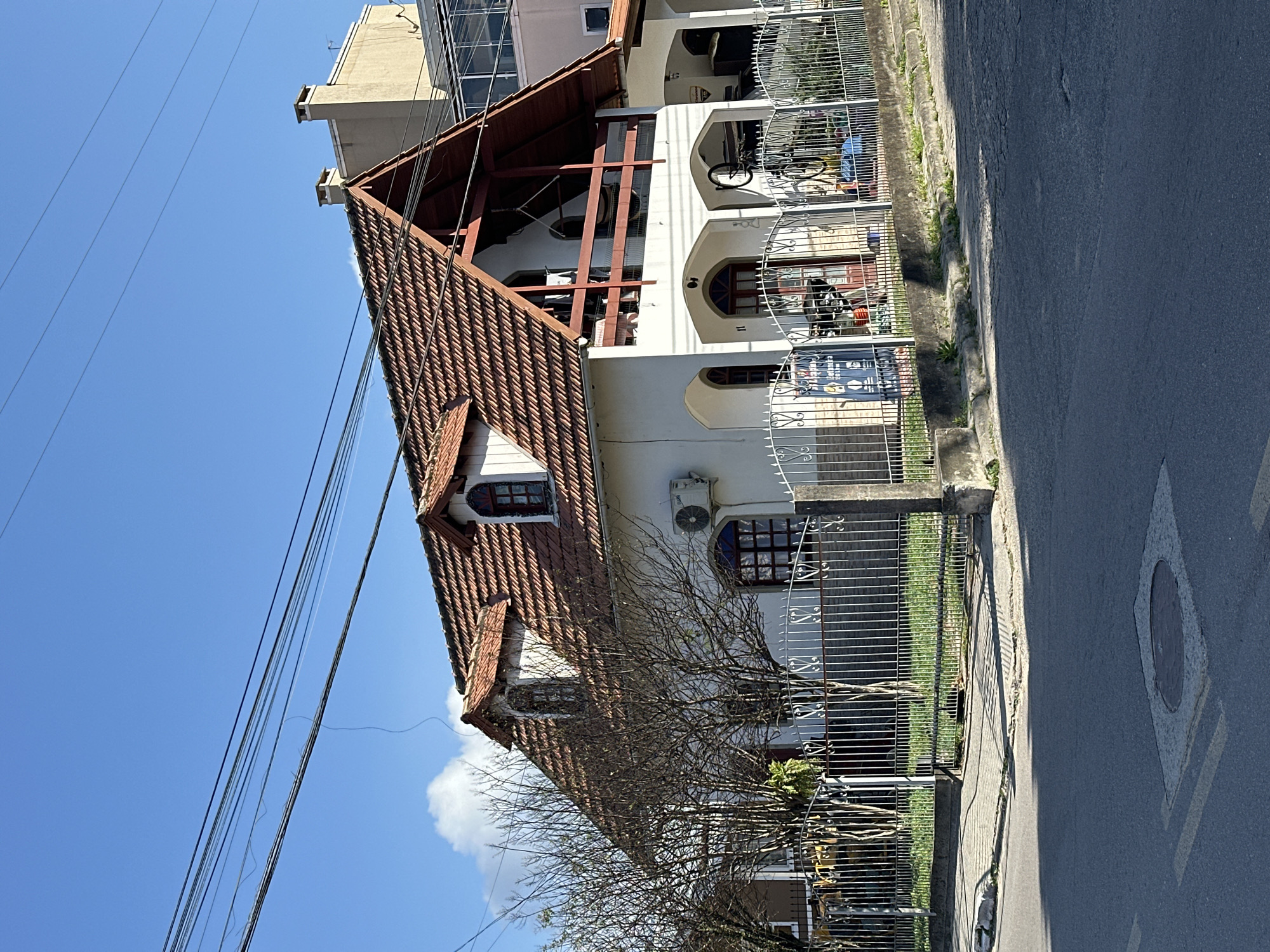 The image shows a two-story white house with a red tile roof, located in Florianópolis, Brazil. The house has a distinctive architectural style with arched windows and a balcony on the second floor. A white metal fence surrounds the property, and a bare tree stands in front of the house. Overhead, power lines are visible against a clear blue sky. The street in the foreground shows asphalt pavement with a manhole cover. The scene appears to be a residential area.