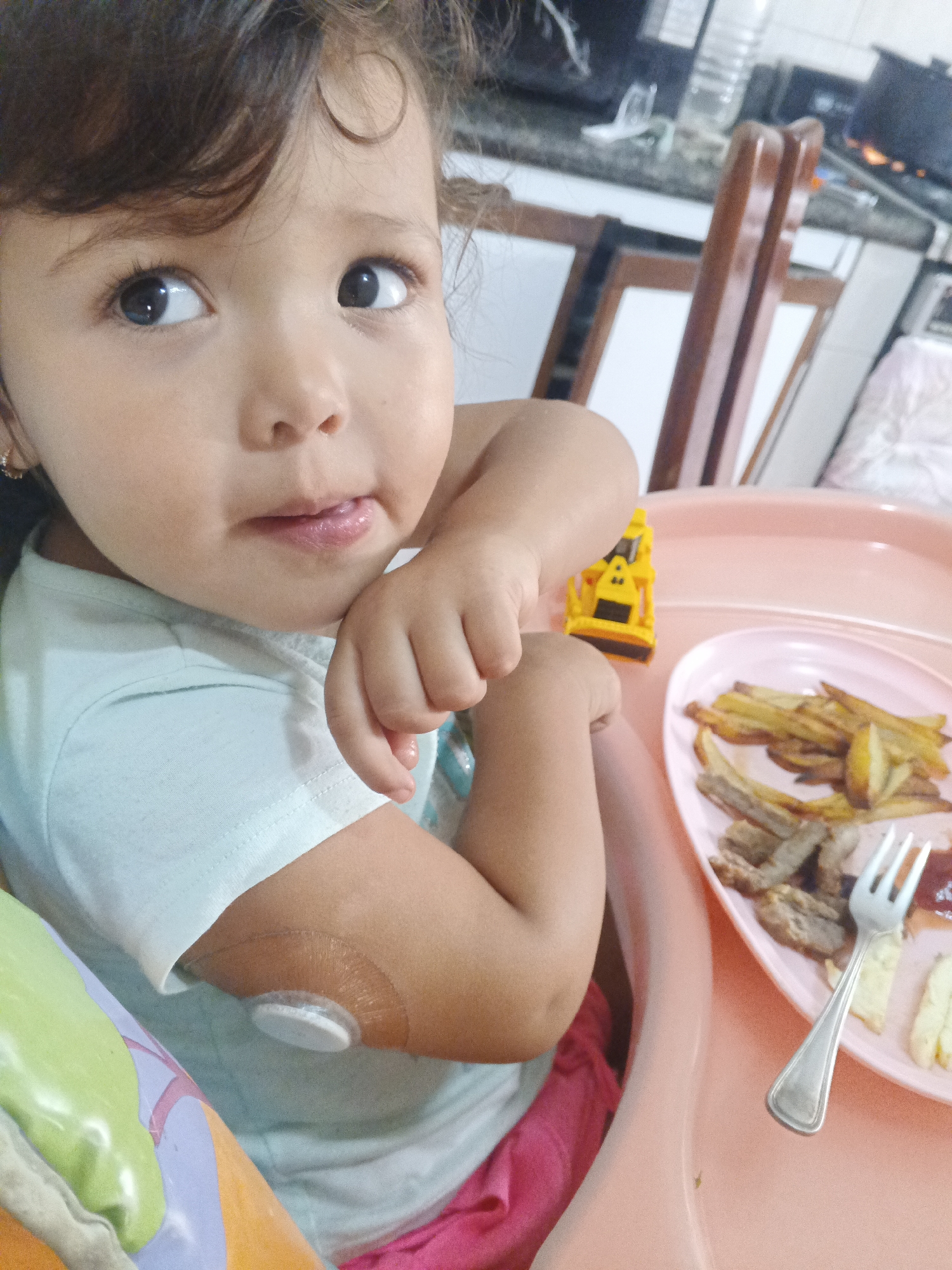 A close-up image shows a young child seated in a pink high chair, looking upward and slightly to the left with a curious or thoughtful expression, and their tongue slightly out. The child has dark, curly hair and dark eyes, and is wearing a light blue short-sleeved t-shirt and partially visible pink shorts. A notable detail is a white, circular medical sensor attached to the child's upper right arm, secured by a clear adhesive film.
On the high chair's tray, to the child's right, rests a pink plate holding a meal of french fries, several pieces of cooked meat, and a small dollop of red sauce, likely ketchup. A silver fork is placed on the plate. Next to the plate, a small yellow toy construction vehicle (an excavator or loader) is visible.
The background suggests an indoor home setting, likely a kitchen or dining area. Elements in the background include wooden chairs with white backrests, a kitchen counter, and what appears to be a stove with a pot on it, along with a water bottle. The lighting suggests it is daytime. No discernible text is visible in the image. The scene is likely a home kitchen or dining area in Caracas, Venezuela.