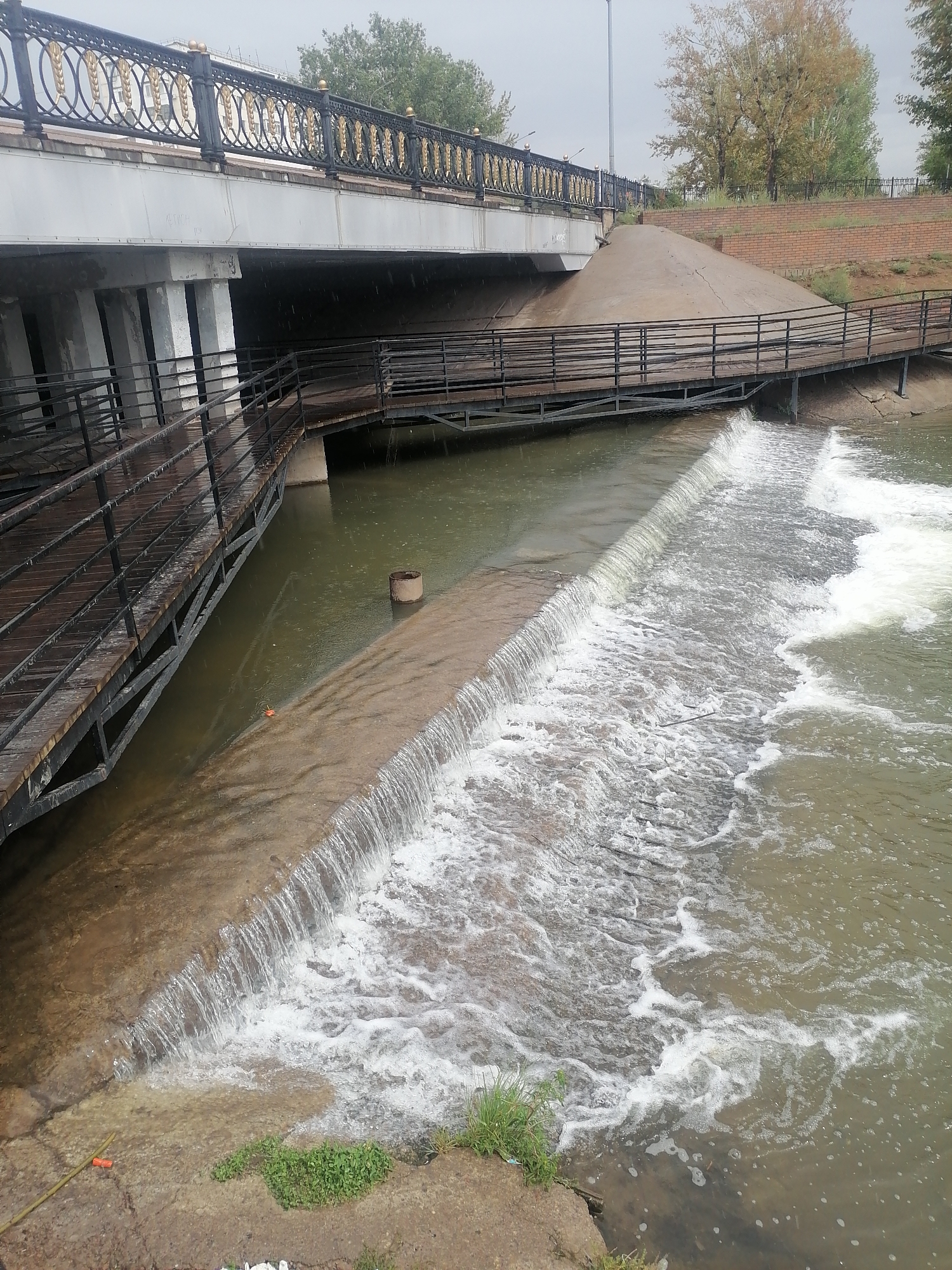 The image depicts a concrete waterfall or weir on a river with wooden boardwalks and metal railings alongside it, under a bridge with decorative railings. The water is choppy and foamy as it cascades over the weir. The riverbanks are visible, with some patches of grass and concrete. The bridge's underside is visible, supported by concrete pillars. Trees with autumn foliage are present in the background, and the sky is overcast. A rusty metal barrel is partially submerged in the water near the weir. There are no people visible in the scene.