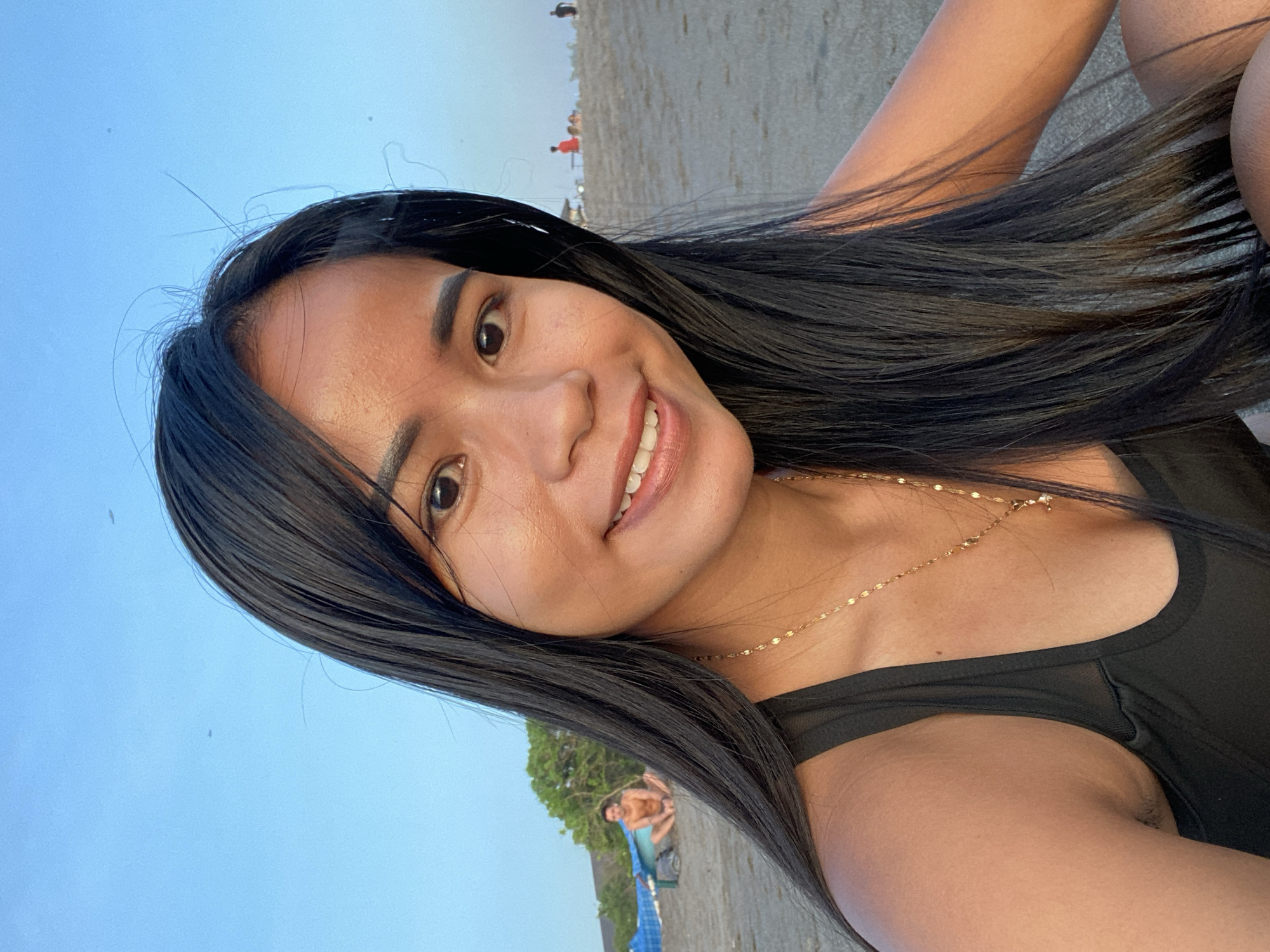 A selfie of a young woman with dark hair, wearing a black top and gold necklace, taken on a beach in North Kuta, Indonesia. She is smiling at the camera. In the background, other beachgoers are visible, along with trees and the ocean. The sky is blue.