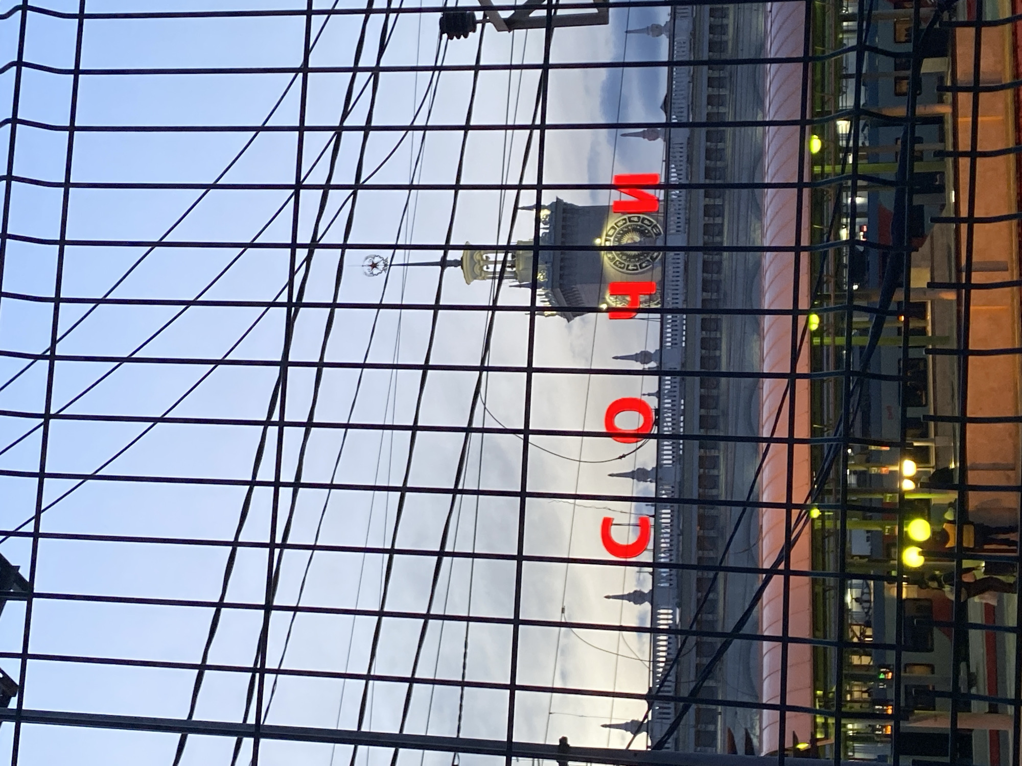 The image shows a partial view of the Sochi Sea Port building in Russia, seen through a metal fence. The building's clock tower and the word "СОЧИ" (Sochi) in red letters are visible. The sky is overcast. Other parts of the building are visible behind the fence, with some lights visible. The fence appears to be in the foreground.