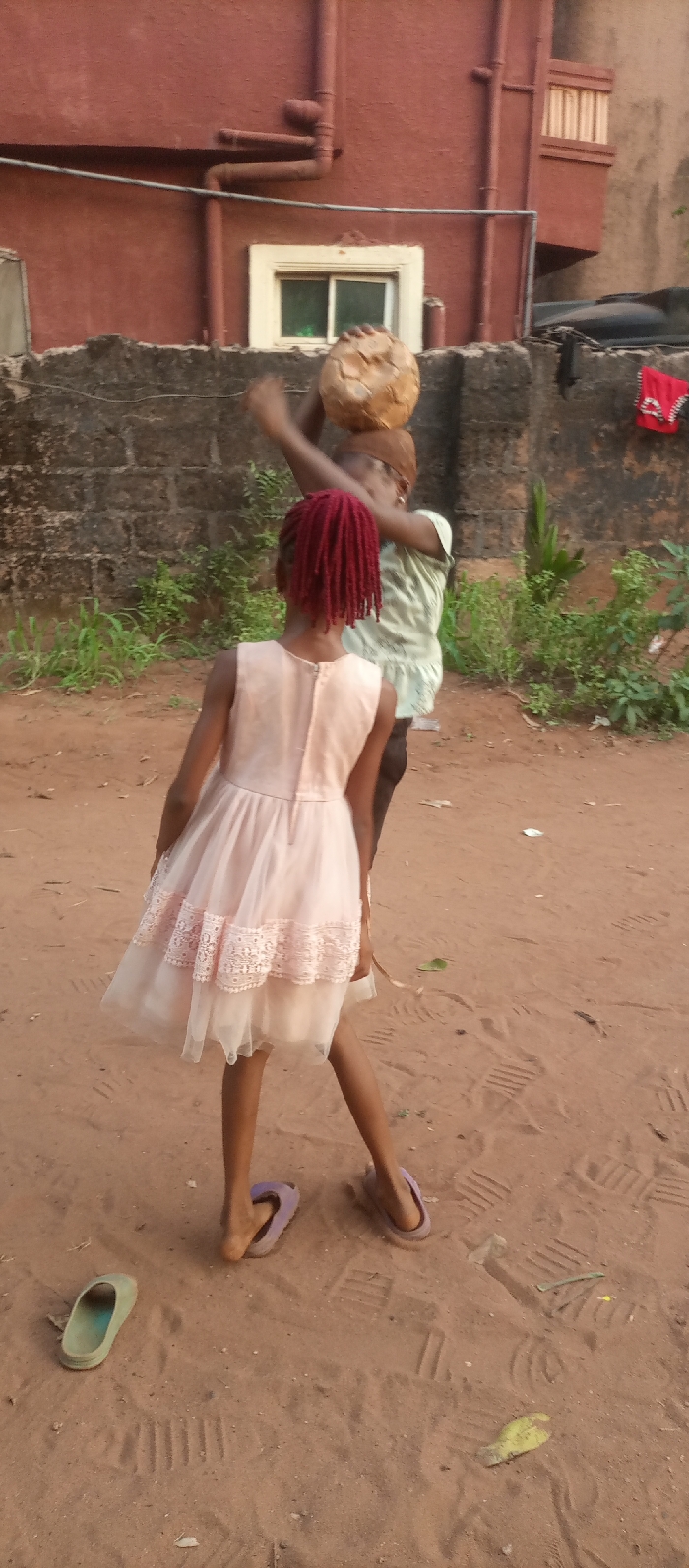 This image, captured in Ogidi, Nigeria, depicts an outdoor scene with two young individuals on a sandy, unpaved surface. In the foreground, facing away from the camera, is a person wearing a light pink sleeveless dress with two tiers of lace trim near the hem, and reddish braided hair. This individual wears light purple slippers, with one foot partially out. To the immediate left on the ground, a single light green slipper rests. Behind and slightly to the right of the foreground individual, a second person is visible, wearing a light-colored top and holding a light brown, spherical object resembling a worn football, raised above their head with both hands. This person's face is partially obscured.
The background features a dark grey concrete block wall, behind which stands a reddish-brown building with a smooth stucco-like finish. A white-framed window with green-tinted panes is centered on this building. Brown drainage pipes are affixed to the building's facade. To the right, a black water storage tank is partially visible behind the wall, and a red piece of cloth or garment is draped over an unseen object or railing. Sparse green vegetation grows along the base of the concrete wall. The sandy ground displays various shoe prints and minor debris.