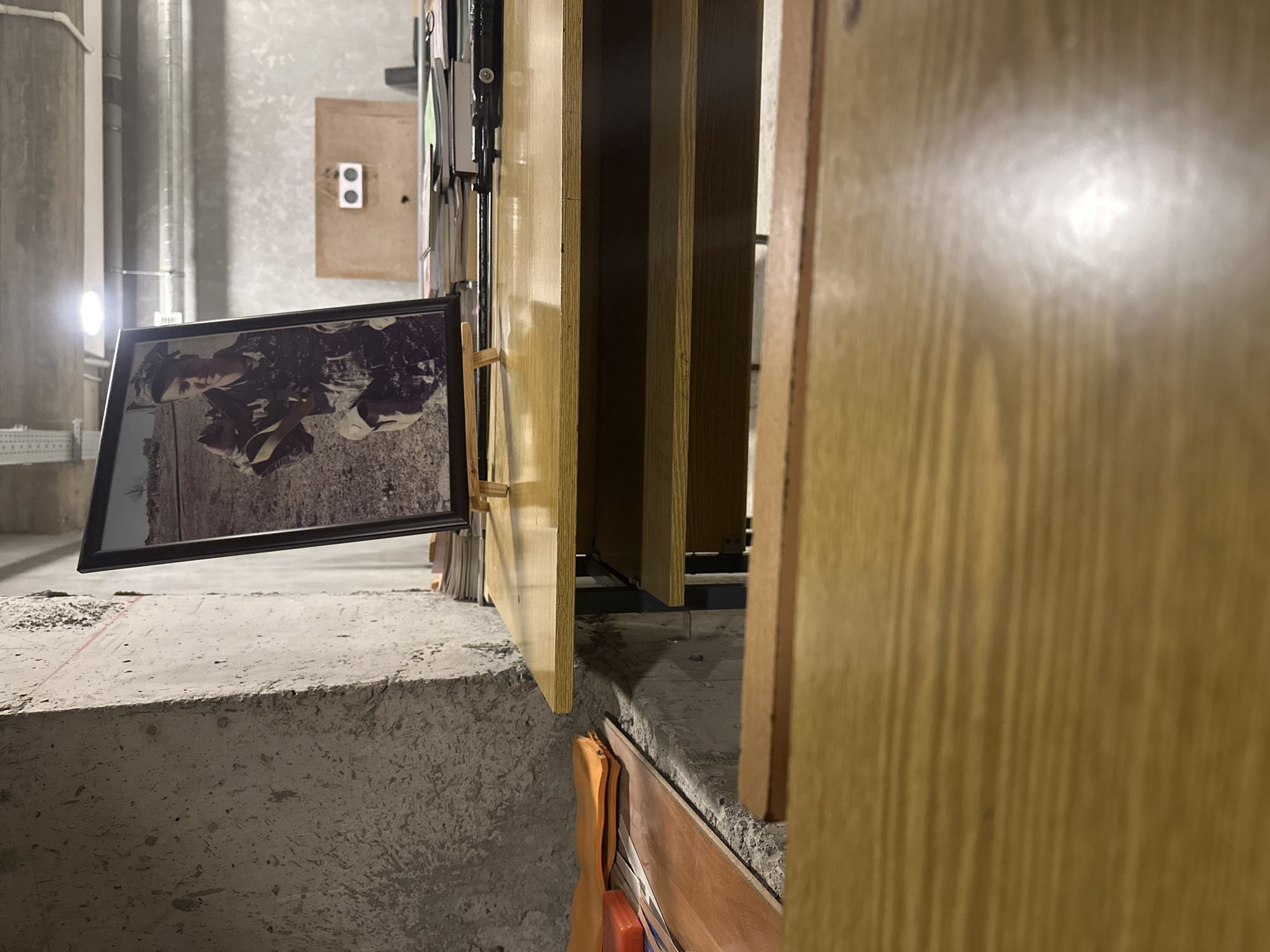 The image shows a close-up of a room under construction. In the foreground are a series of vertical wooden panels. A framed photograph rests on the edge of a concrete block. The photograph's image is of a person in dark clothing. Behind the photograph and wooden panels are pipes and a rectangular plate.