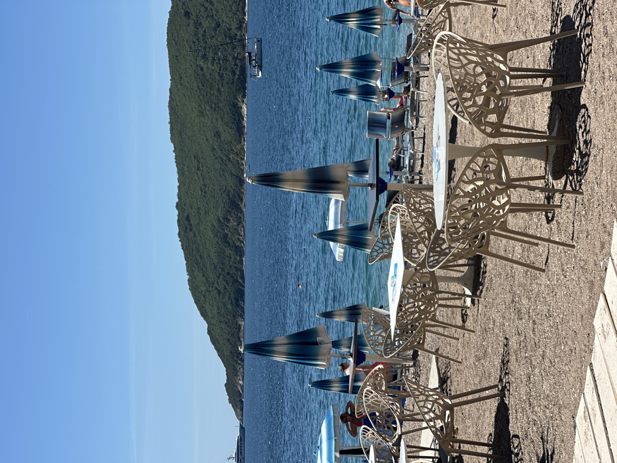 The image captures a coastal scene, likely a beachside cafe or resort. There are rows of open-air tables and chairs with intricate webbed designs set upon a gravelly surface alongside a white stone pathway. Blue and white striped beach umbrellas provide shade to seated individuals. The sea extends to the left of the frame, and there's a cliff covered in lush green vegetation lining the coastline. Overhead, a cable car system is visible, suggesting a mountainous terrain in close proximity to the coast. Clear blue skies are visible in the upper left corner, indicating a sunny day. Based on the architectural style and landscape features, this scene is likely located in Budva, Montenegro.