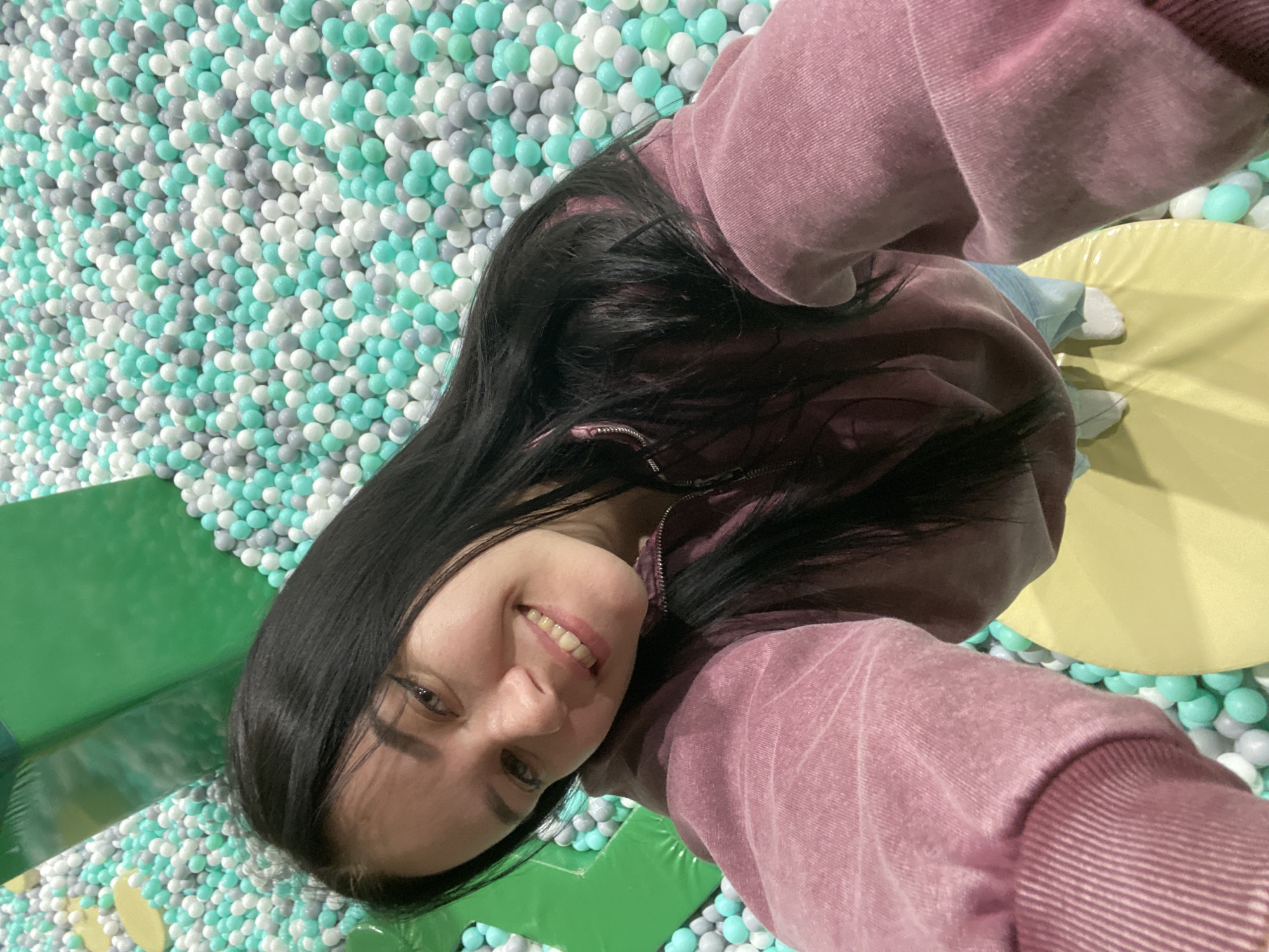 A woman with dark hair is in a ball pit, possibly at a family entertainment center. The pit contains balls in shades of white, gray, and teal. The woman is wearing a pink hooded sweatshirt and is smiling at the camera. There are padded mats in green and yellow visible near the ball pit. The location is likely in Perm, Russia.