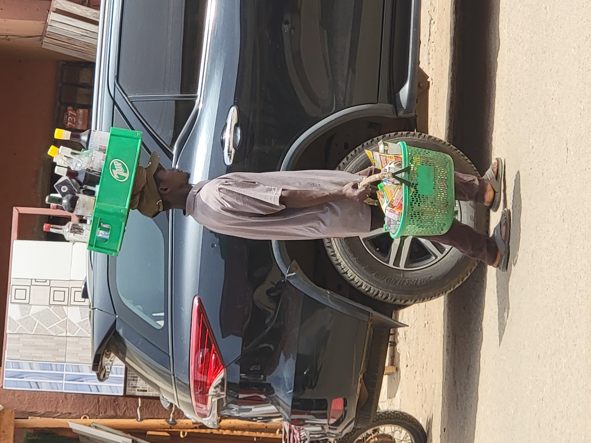This image, set in Maiburiji, Nigeria, captures a male street vendor carrying merchandise under bright daytime sunlight. The vendor is seen standing outdoors, next to a dark-colored SUV parked on a road or path.
On his head, the vendor expertly balances a rectangular green plastic crate filled with numerous small bottles, likely spirits or energy drinks, identifiable by their various yellow, red, and white caps. One bottle clearly shows the text "737" on its label. The green crate itself features a distinct white logo on its side. In his right hand, he carries a green plastic basket brimming with an assortment of packaged snacks and other small items.
The vendor is dressed in a light brown or grey long-sleeved shirt and dark brown trousers, with flip-flops on his feet. The background shows parts of the black SUV, including a section of its rear wheel with "BRIDGESTONE" visible on the tire, a red taillight, and a chrome door handle. Behind the vehicle, a light-colored wall is present, and to the far left, stacked building materials, possibly tiles, are visible. A portion of a bicycle wheel can be seen in the bottom left of the frame. The scene portrays a typical street vending activity in the region.