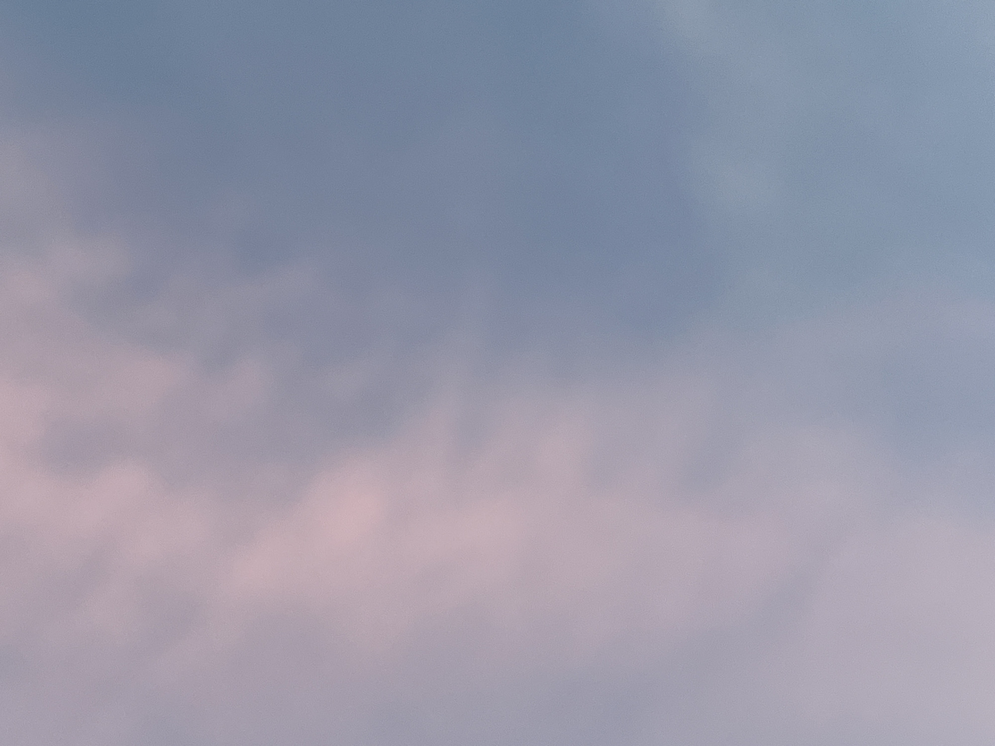 This is an abstract, out-of-focus image of the sky. The colors are muted, with soft pinkish-purple clouds on the left blending into a pale blue sky on the right. The image appears to be taken from a low angle, looking upwards. There are no discernible objects, people, or specific landmarks visible that would allow for identification of a location within Saint Petersburg, Russia. The texture is grainy, suggesting it might be a photograph with some noise or a digital rendering with a simulated texture.