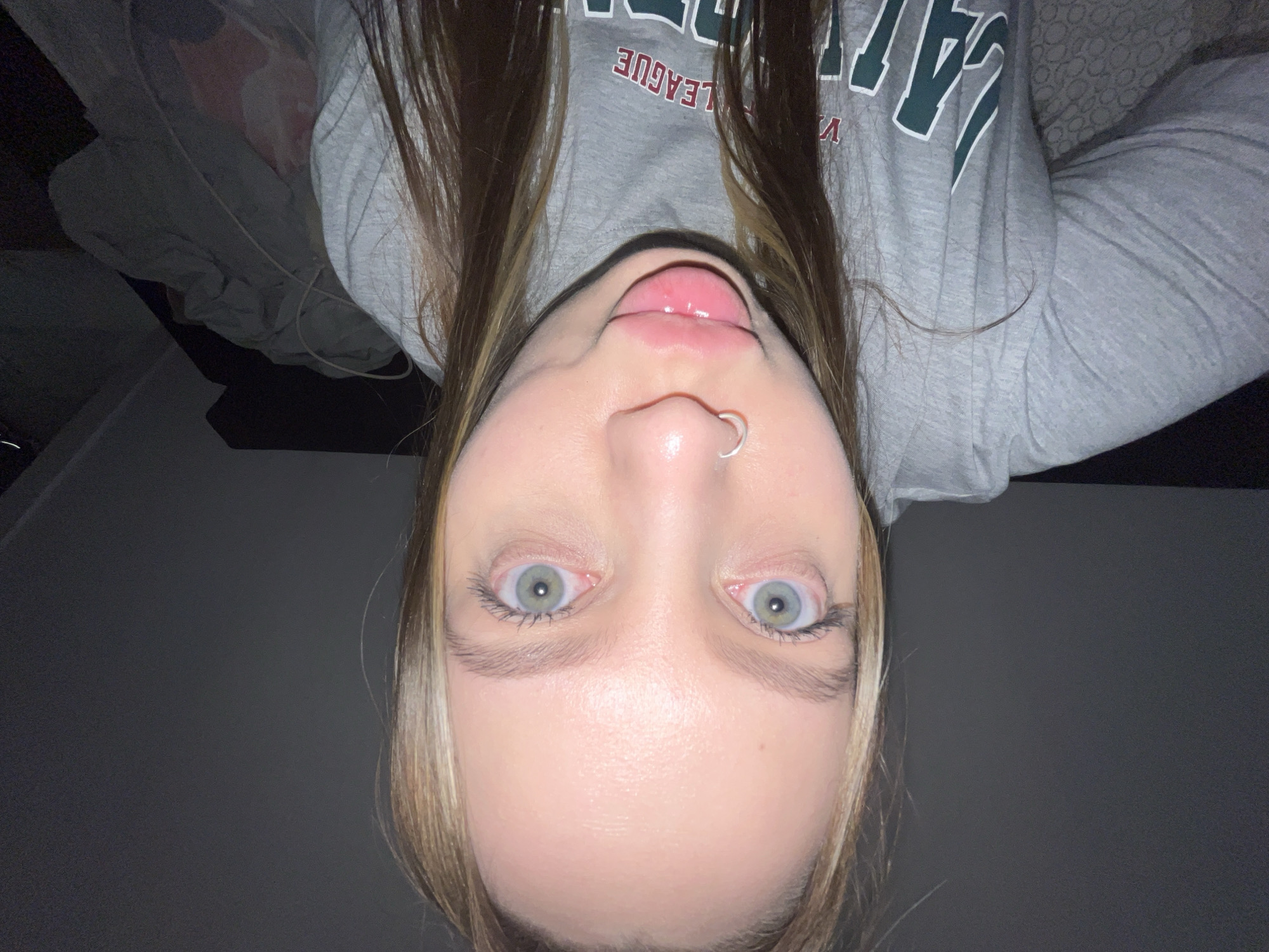 A wide-angle, close-up photograph depicts a young adult female indoors in Chaikovsky, Russia. The subject has light skin, light green-blue eyes with dilated pupils, and light brown hair with blonde highlights. She features a silver-colored hoop piercing in her left nostril. Her lips are pursed forward in an exaggerated pose, appearing glossy. She is wearing a grey long-sleeved top with a visible green and white graphic print on the chest, partially reading "V" and "LEAGUE". The background behind the subject is dark, with a patterned surface, possibly bedding, visible on the lower left. The lighting is dim, suggesting an indoor setting illuminated primarily from the front.