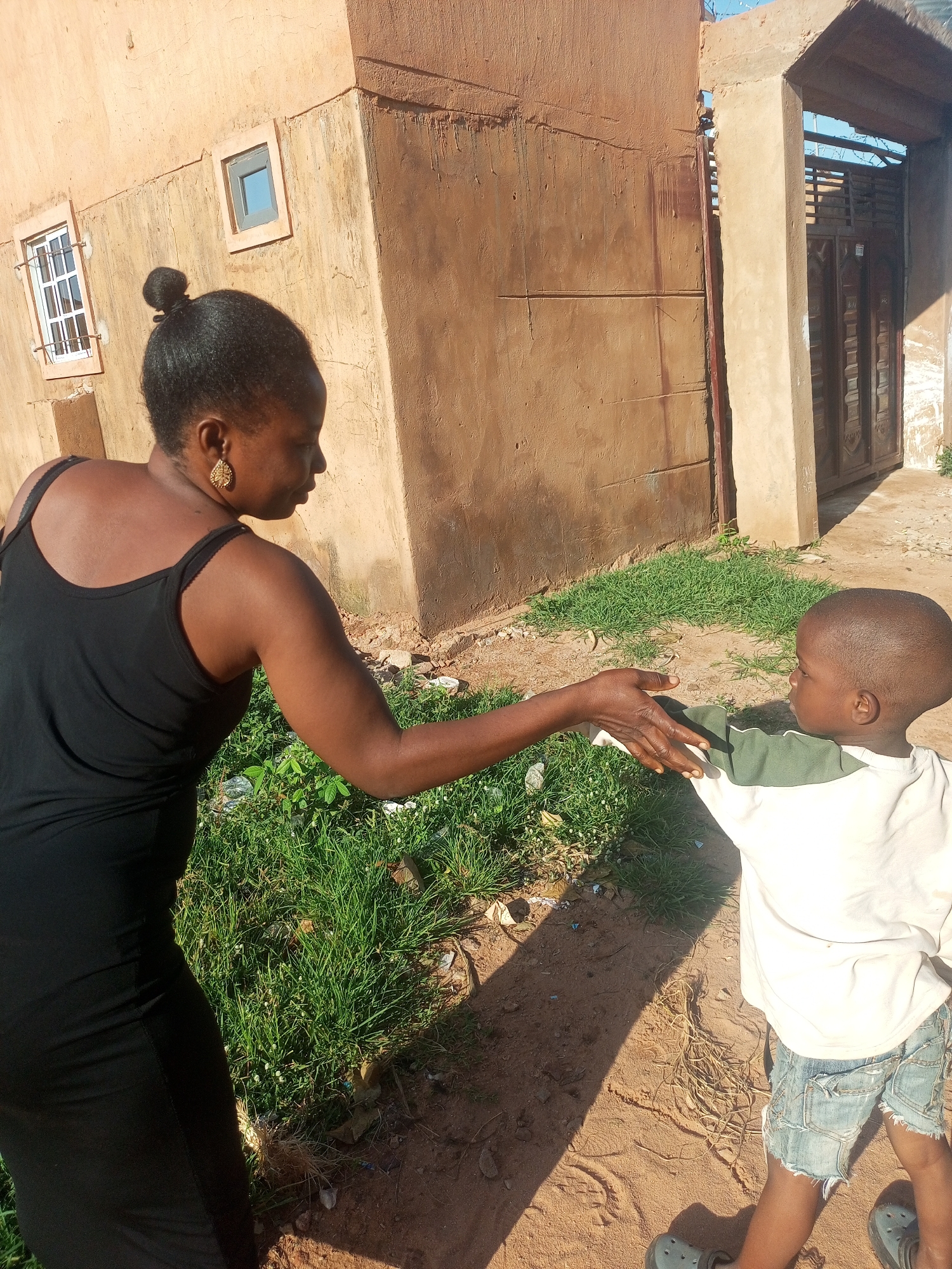 A woman in a black dress and a young boy in a white and green shirt and denim shorts are shaking hands outside a building in Tempure, Nigeria. The building is made of clay and has two windows. The ground is dirt and grass, and there is a shadow cast from the building. The sky is blue.
