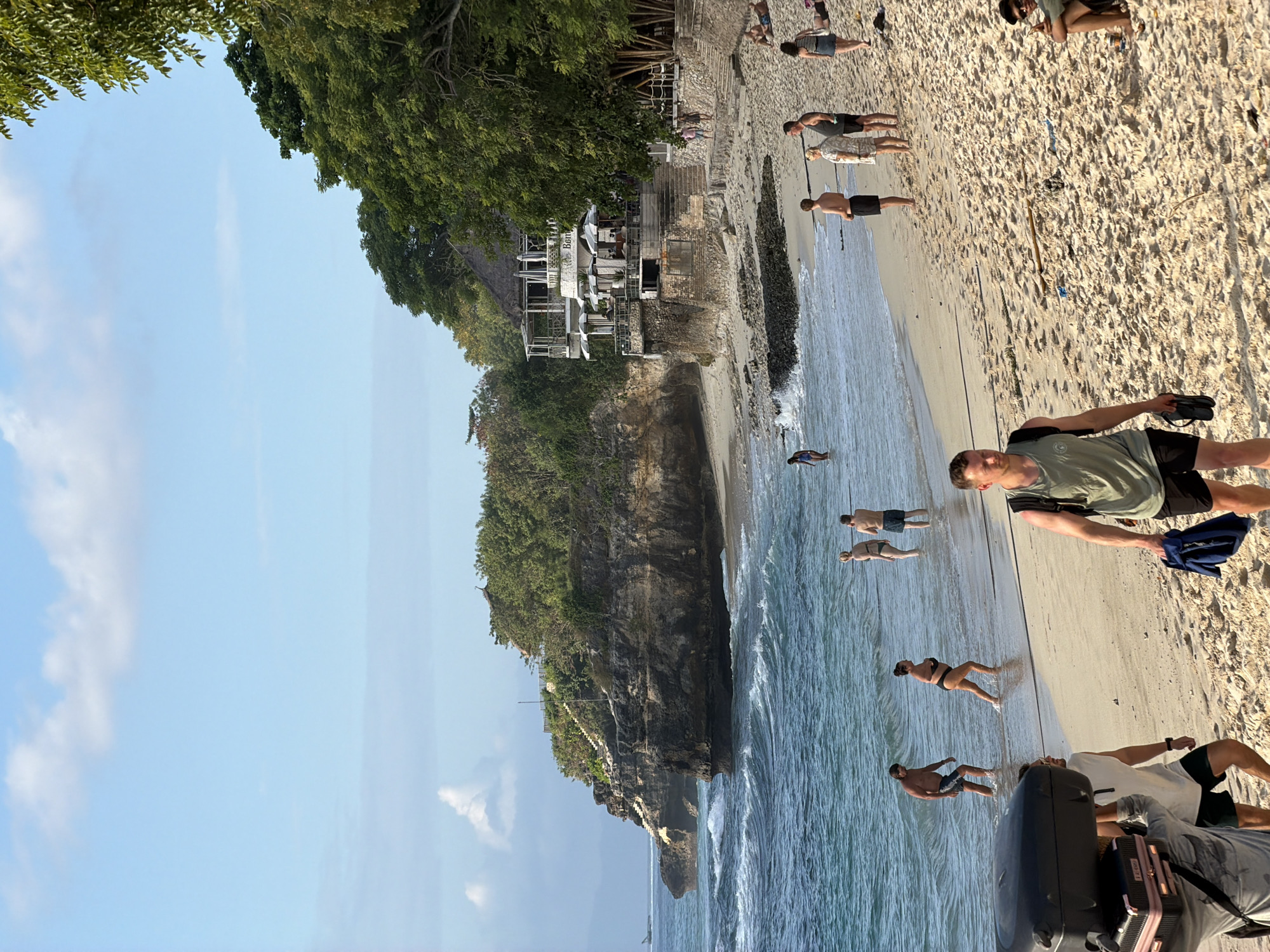 The image depicts a bustling beach scene in Nusa Penida, Indonesia, likely near the popular cliffside restaurant, "The Baan". The foreground is dominated by a sandy beach, with several individuals visible. A man in the lower right foreground, wearing a green tank top and black shorts, walks towards the viewer, holding a pair of flip-flops. To his left, a person is seated on the sand. Further up the beach, a group of people are standing near the water's edge, appearing to be watching the waves or socializing. Several individuals are in the shallow water, some wading, while others are further out. The middle ground features the ocean with moderate waves and a prominent cliff face on the left, topped with lush green trees. A multi-level building, likely the aforementioned restaurant, is perched on the cliff, with umbrellas and seating areas visible. Steps lead up the cliff towards the building. The background shows a clear blue sky with scattered clouds, and the distant horizon line. The overall impression is one of a vibrant, sunlit day at a popular tourist destination.