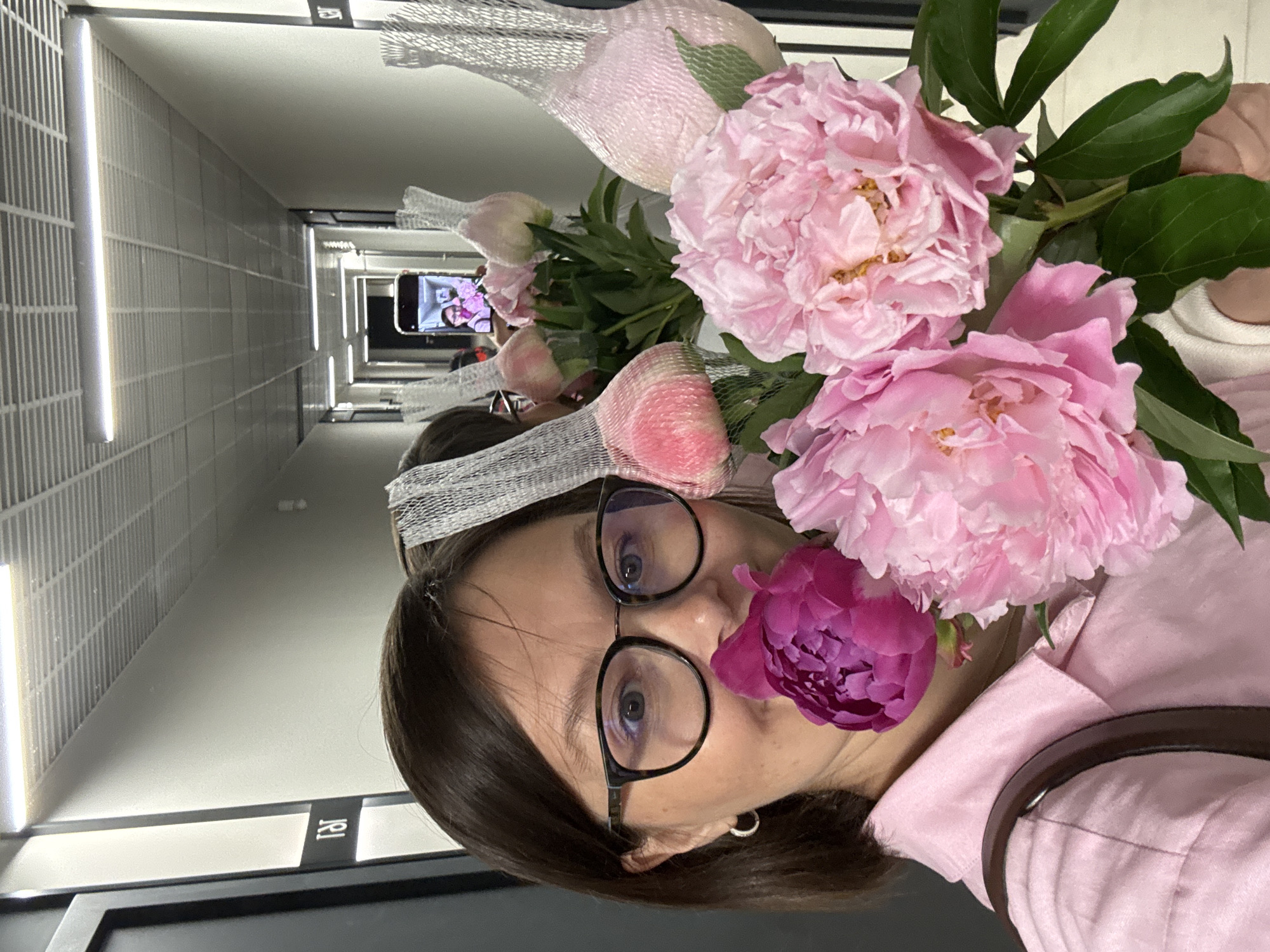 A woman with dark brown hair, wearing glasses and a pink shirt, holds a bouquet of peonies in a hallway. She is taking a selfie with her phone, which is visible in her hand and shows a reflection of herself and the flowers. The hallway has white walls, a tiled ceiling with fluorescent lights, and doors on either side. Some of the peonies are wrapped in white netting.